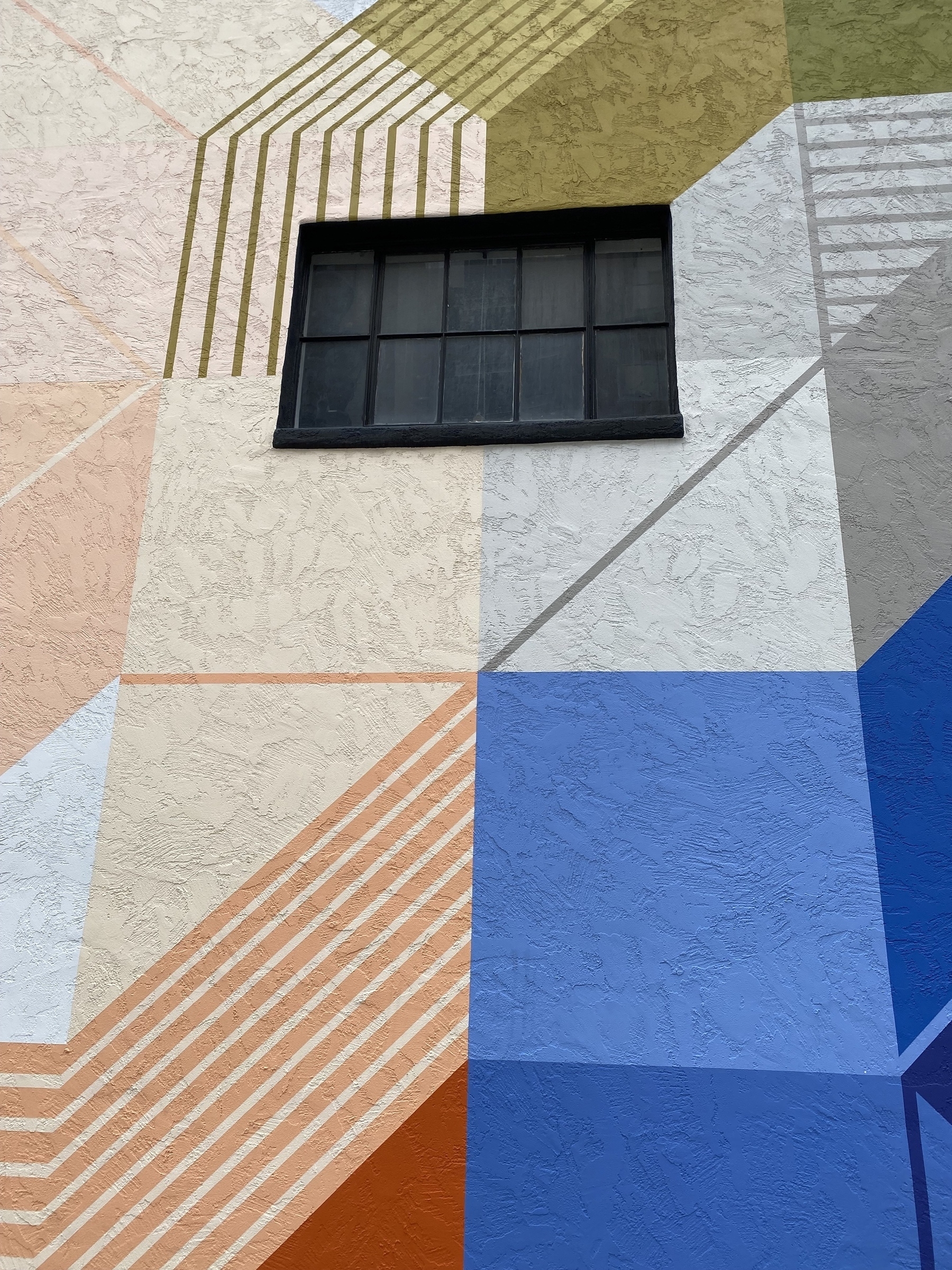Mural on the side of a building of geometric shapes in various colors, a single dark window centered above.
