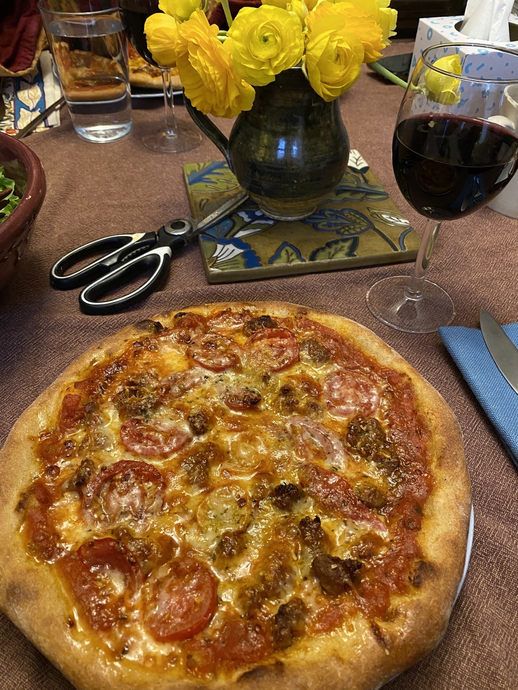 Small pizza on a plate next to a glass of wine, a vase of yellow roses, and a pair of kitchen scissors.