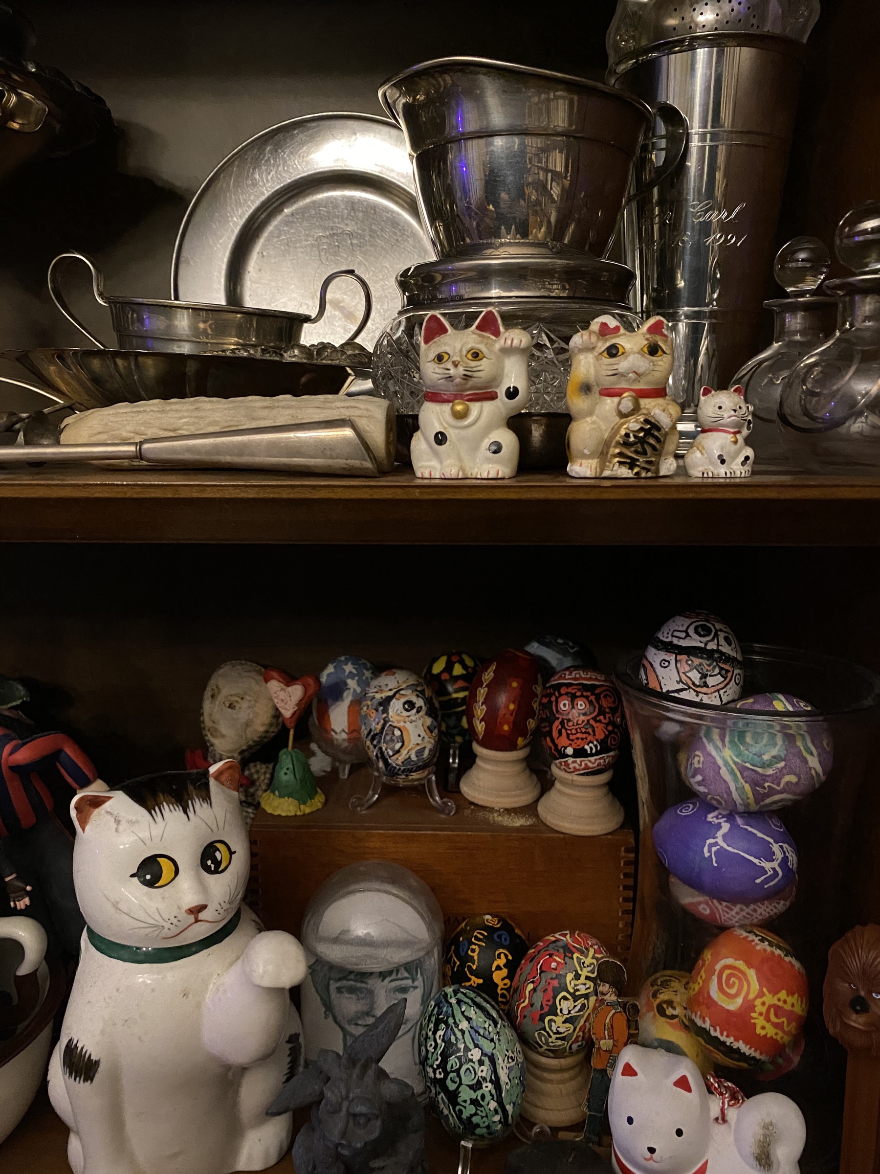 Close up of a cabinet with silver cups, pysanky eggs, and lucky cat figurines.