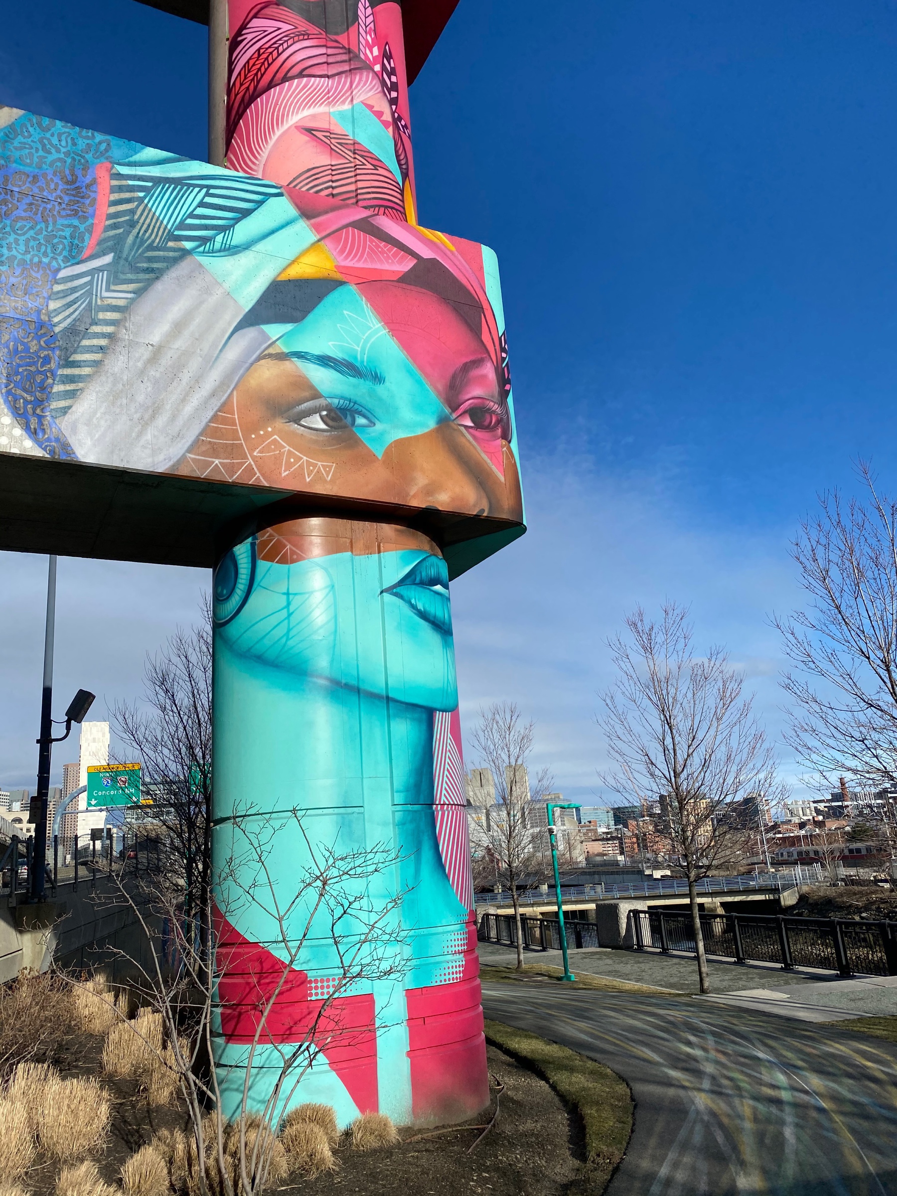 Mural of a woman's face in cyan and magenta on the column of a highway overpass.