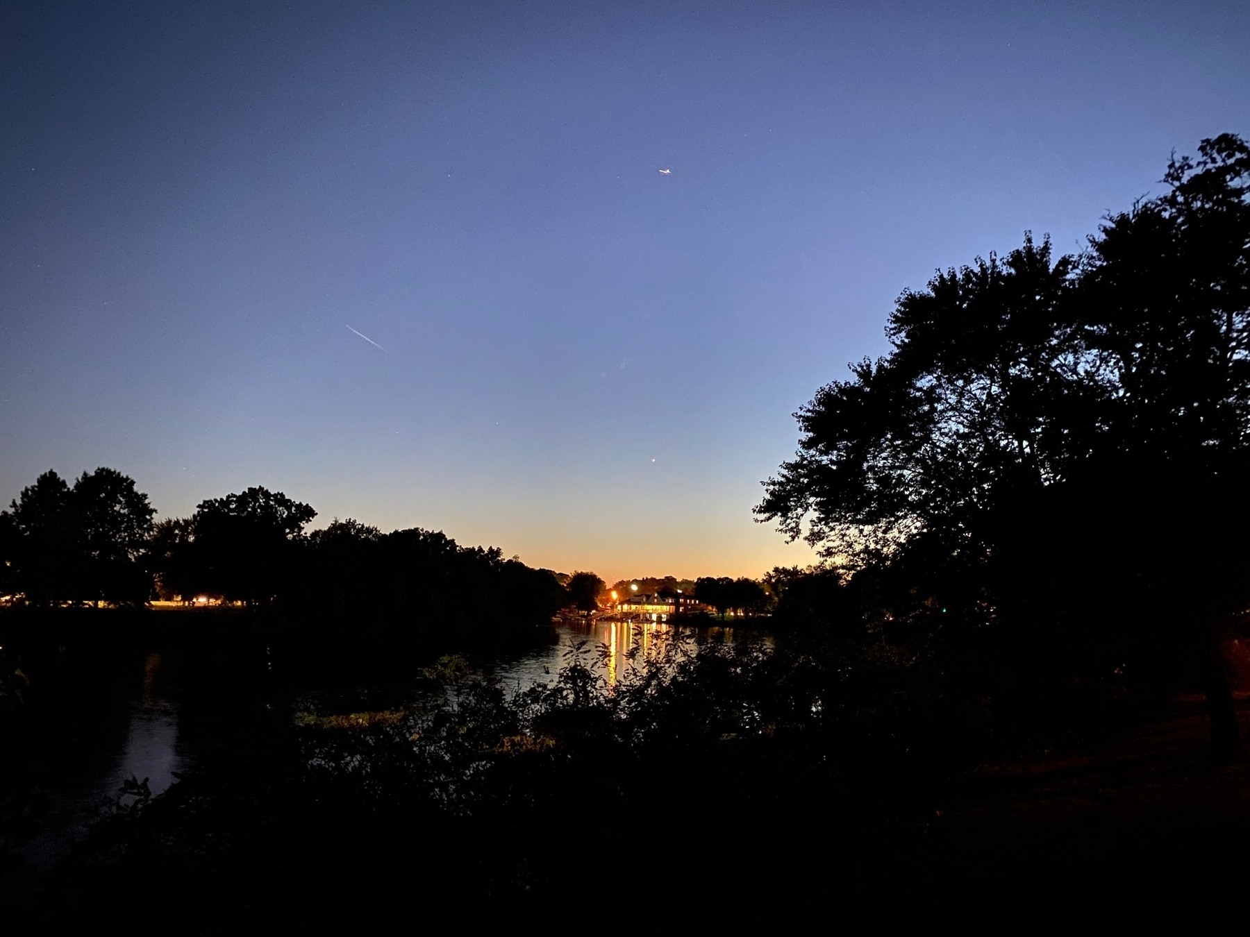 View of a river just after sunset, with a brightly lit boathouse on the far shore.