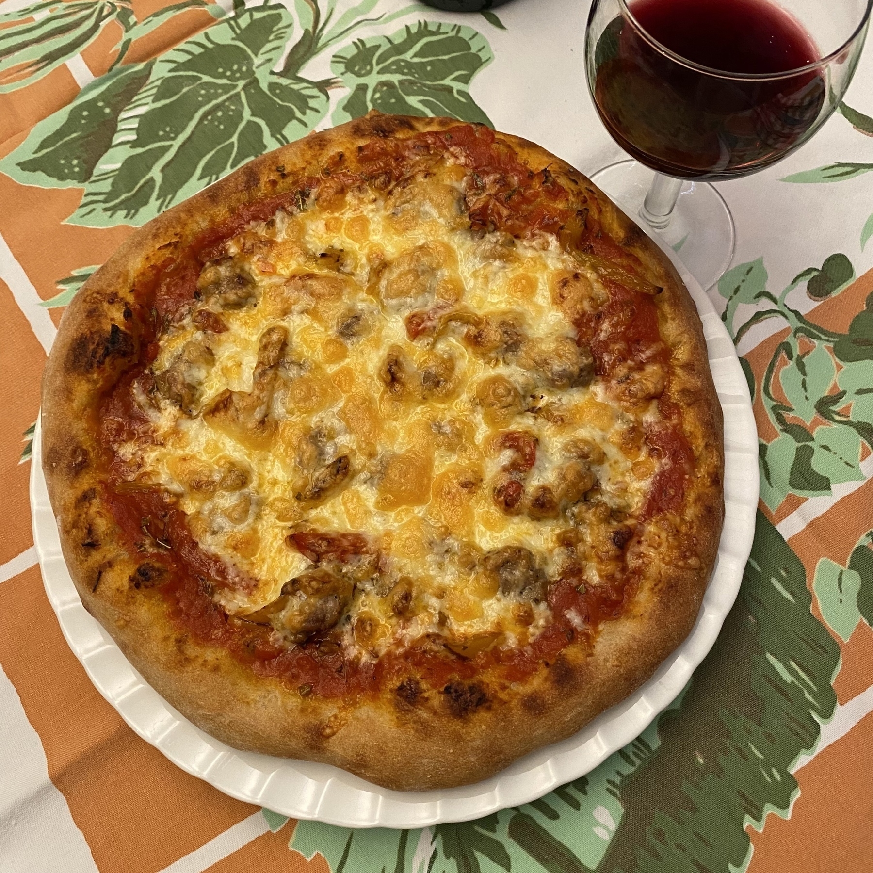 Small pizza on a white plate, sitting on a table cloth next to a glass of red wine.