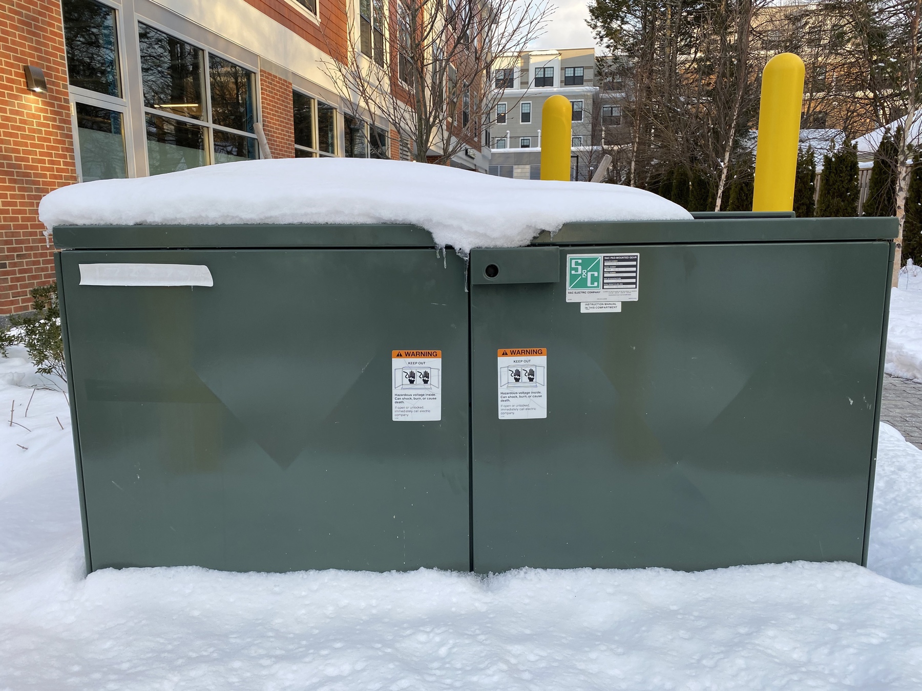 View of a power box behind a brick building, covered in snow.