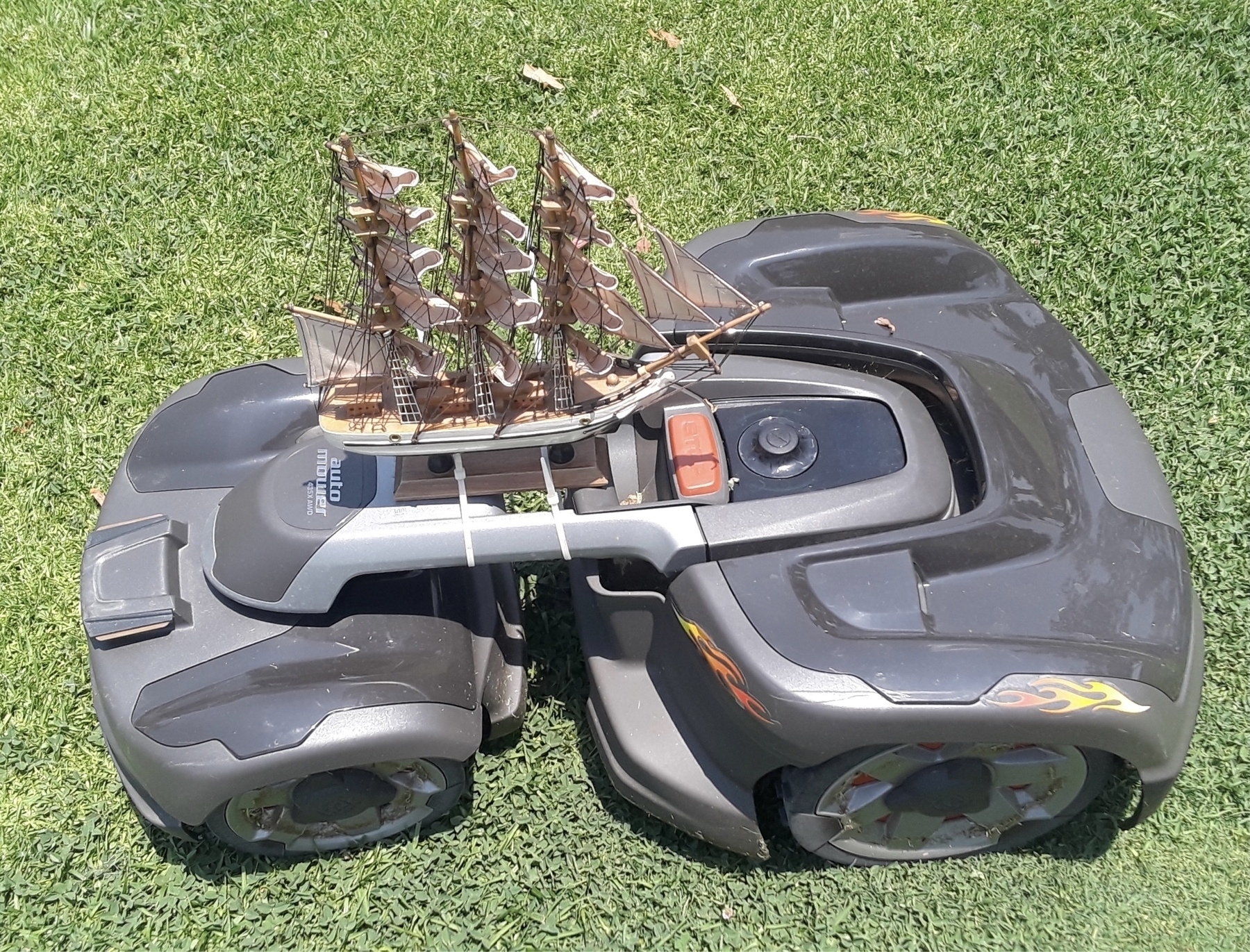 Close up view of a gray lawn mowing robot with a small model sailing ship attached to the top.