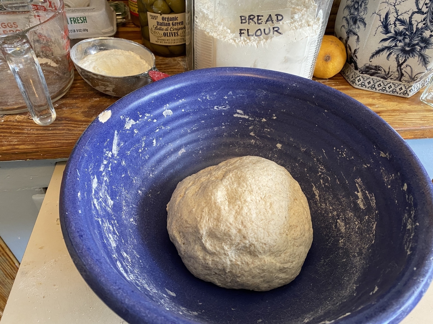 Close up of a dough ball in a blue bowl, with a jar of flour and a lemon on the shelf behind.
