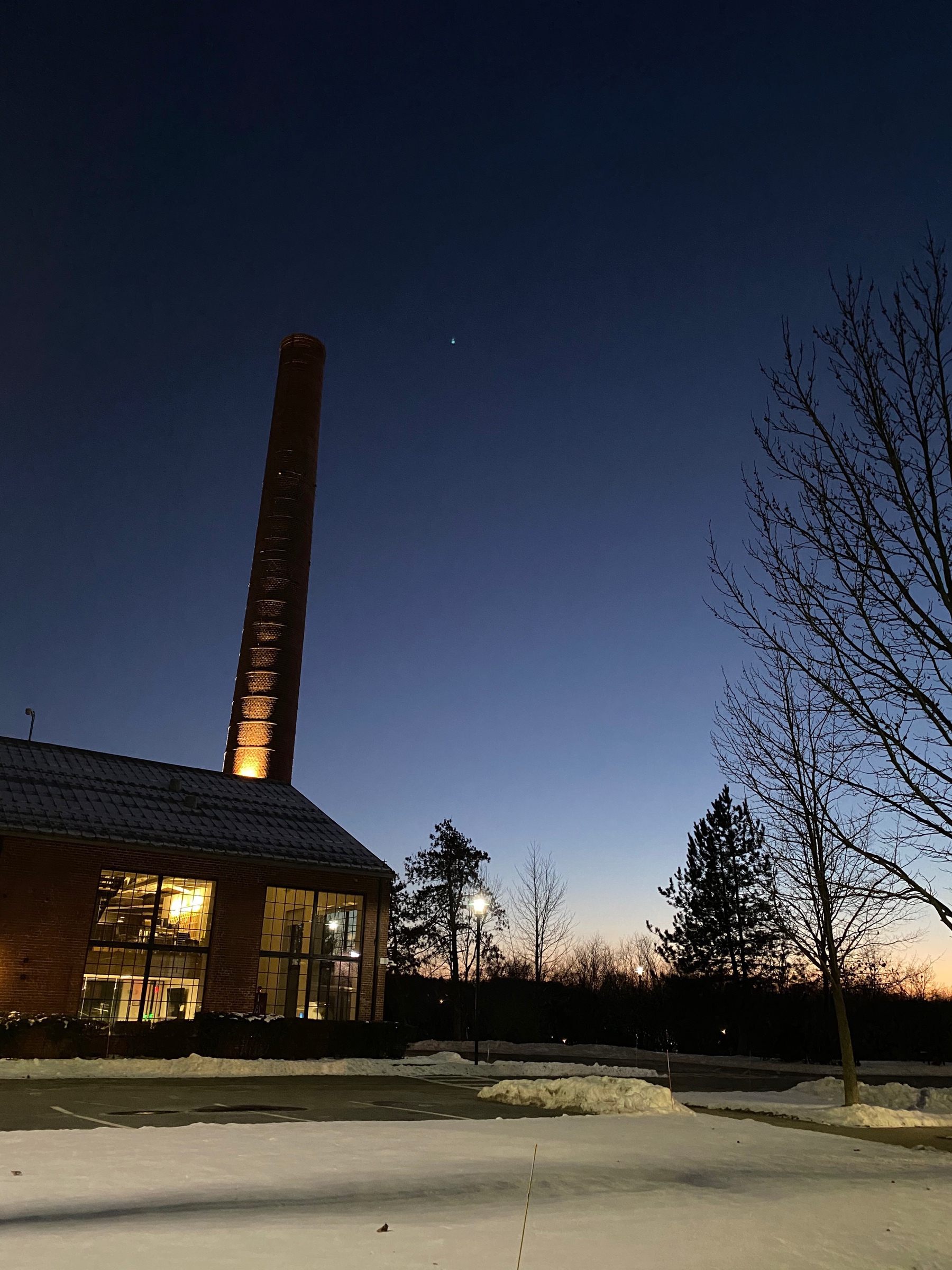 View shortly after sunset with the horizon still pink and Venus in the sky, trees and an illuminated building with a tall smokestack in the forground.