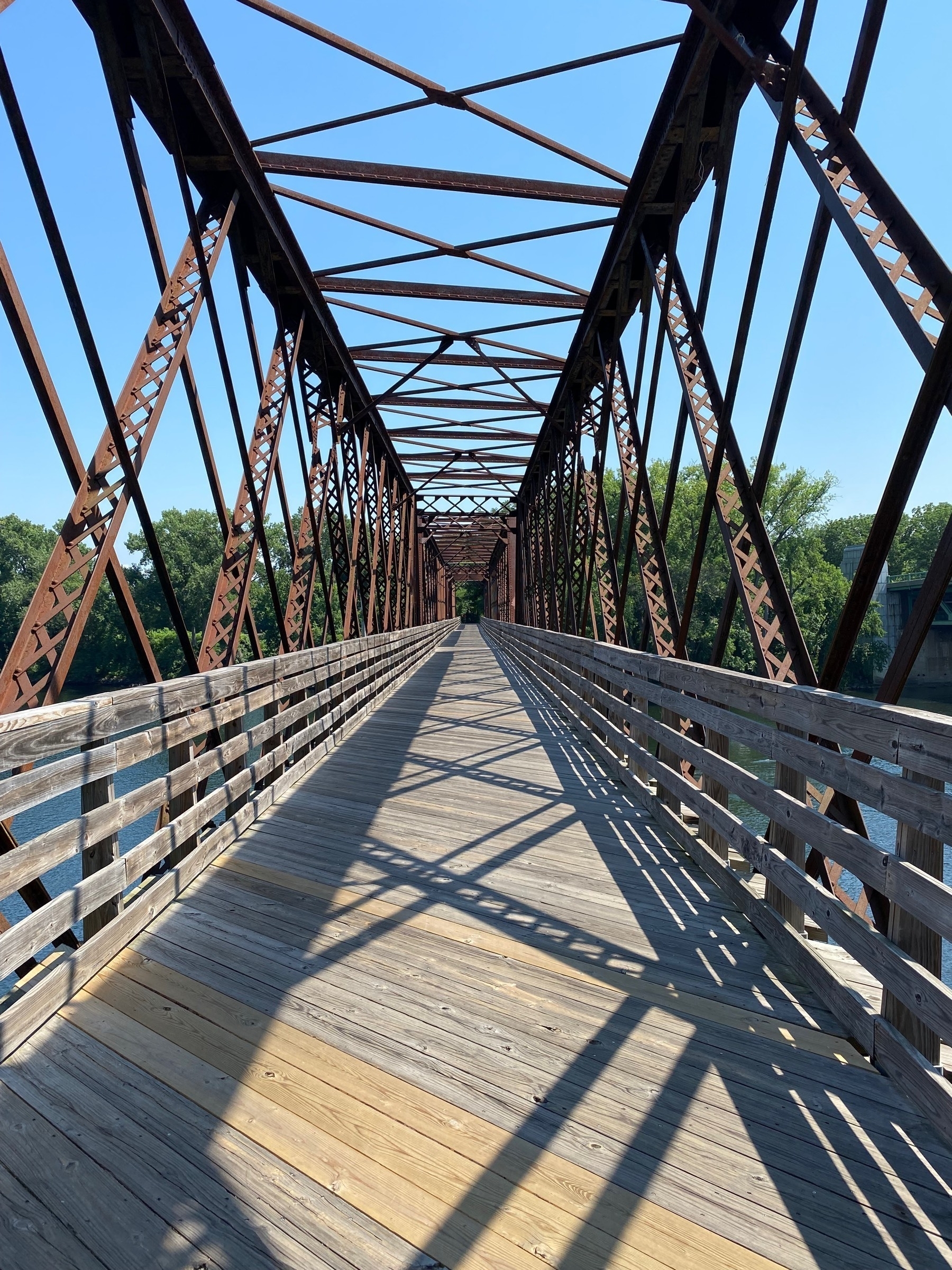 View of a bike path crossing the Connecticut River over an old railway bridge, the lattice of girders above casts shadows on the wooden boards below.