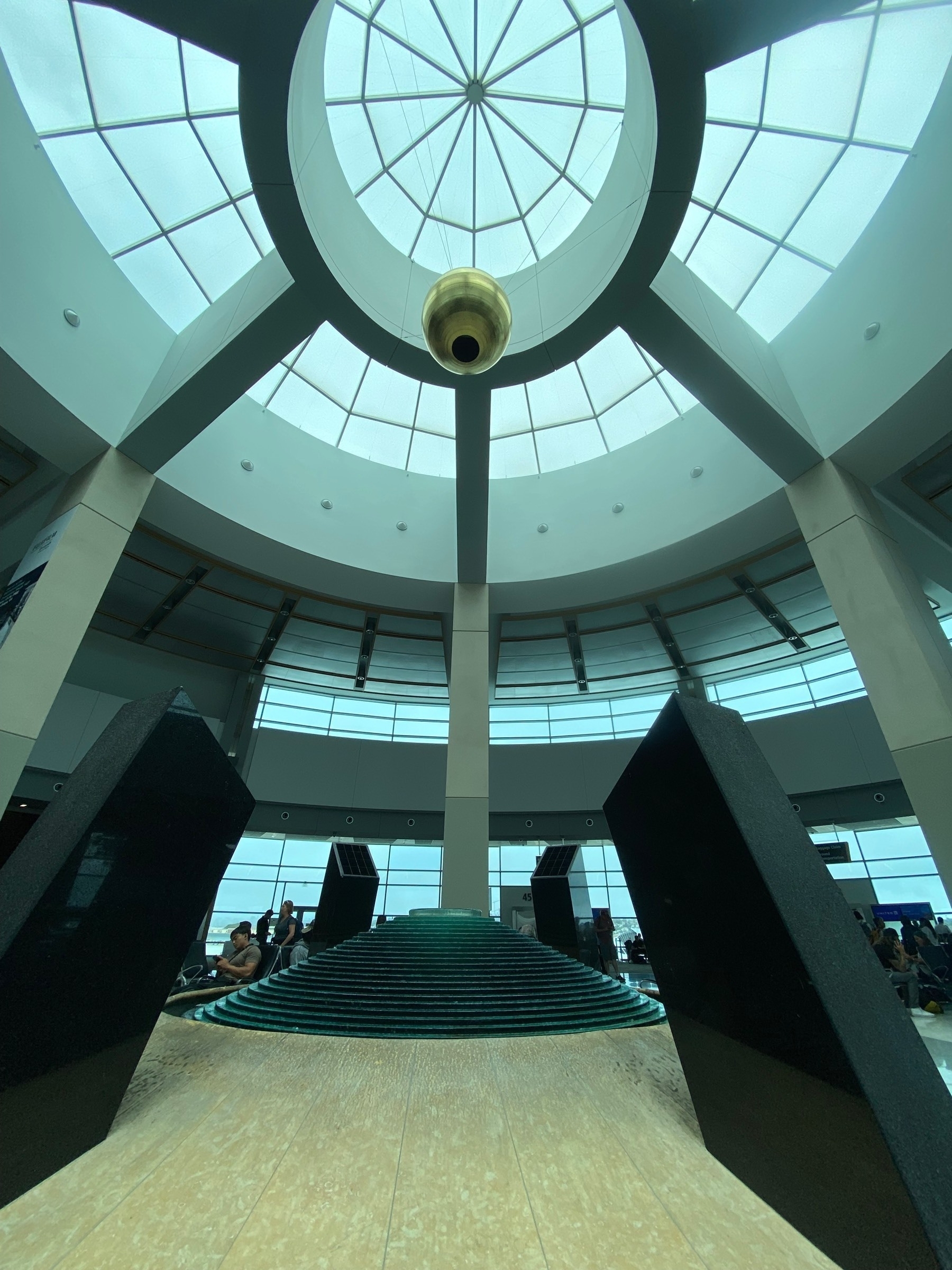 A circular fountain surrounded by obelisk below a golden ball hanging from a domed skylight.