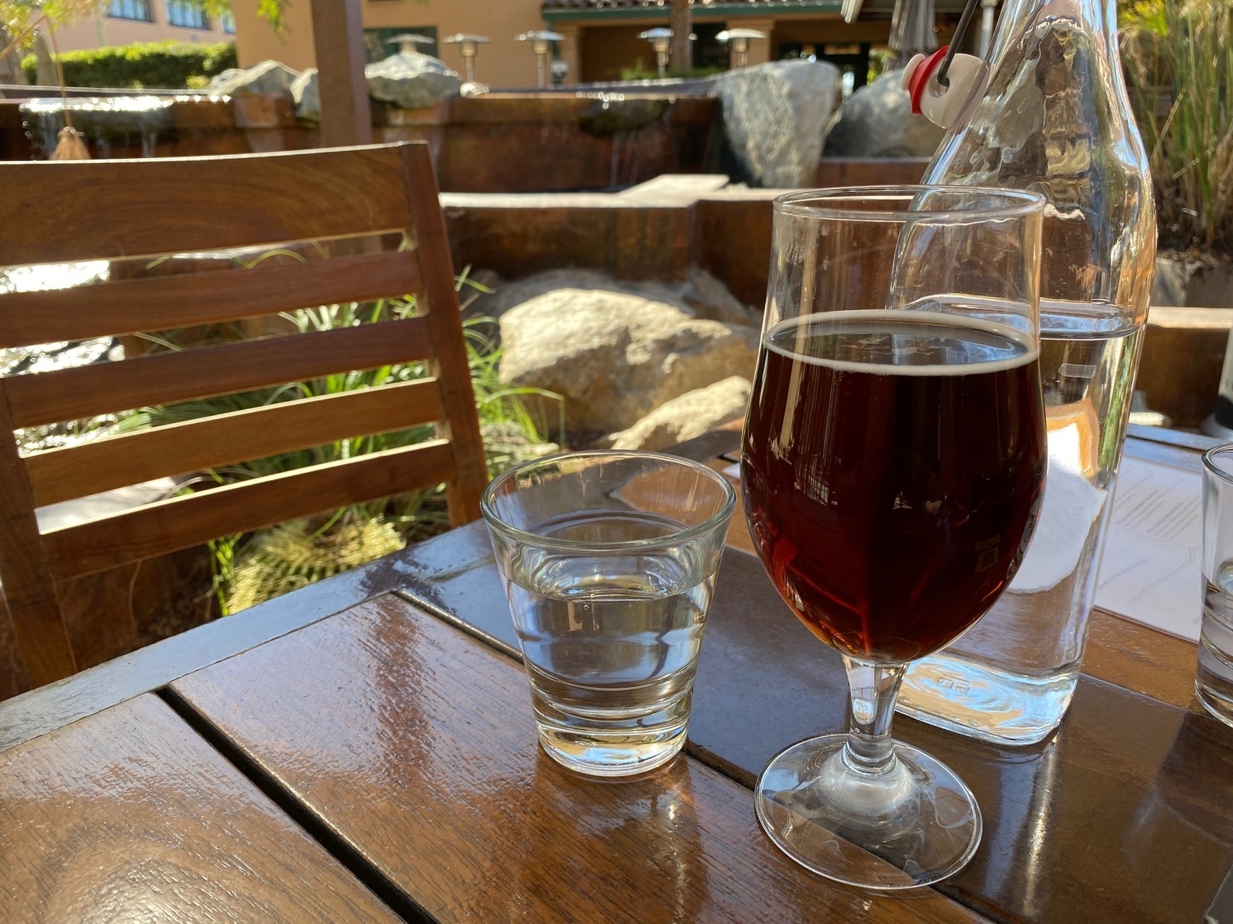 A glass of beer on a wooden table with a small fountain in the background.