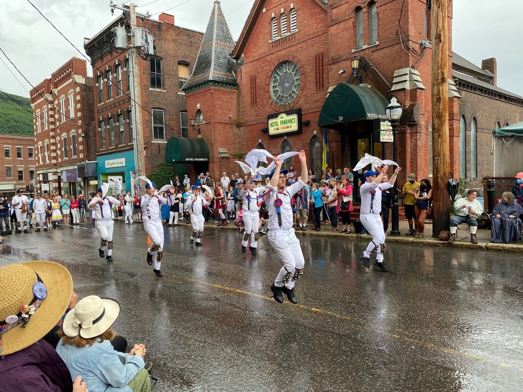 Morris dancers waving handkerchiefs while dancing on a wet street in front of brick buildings.