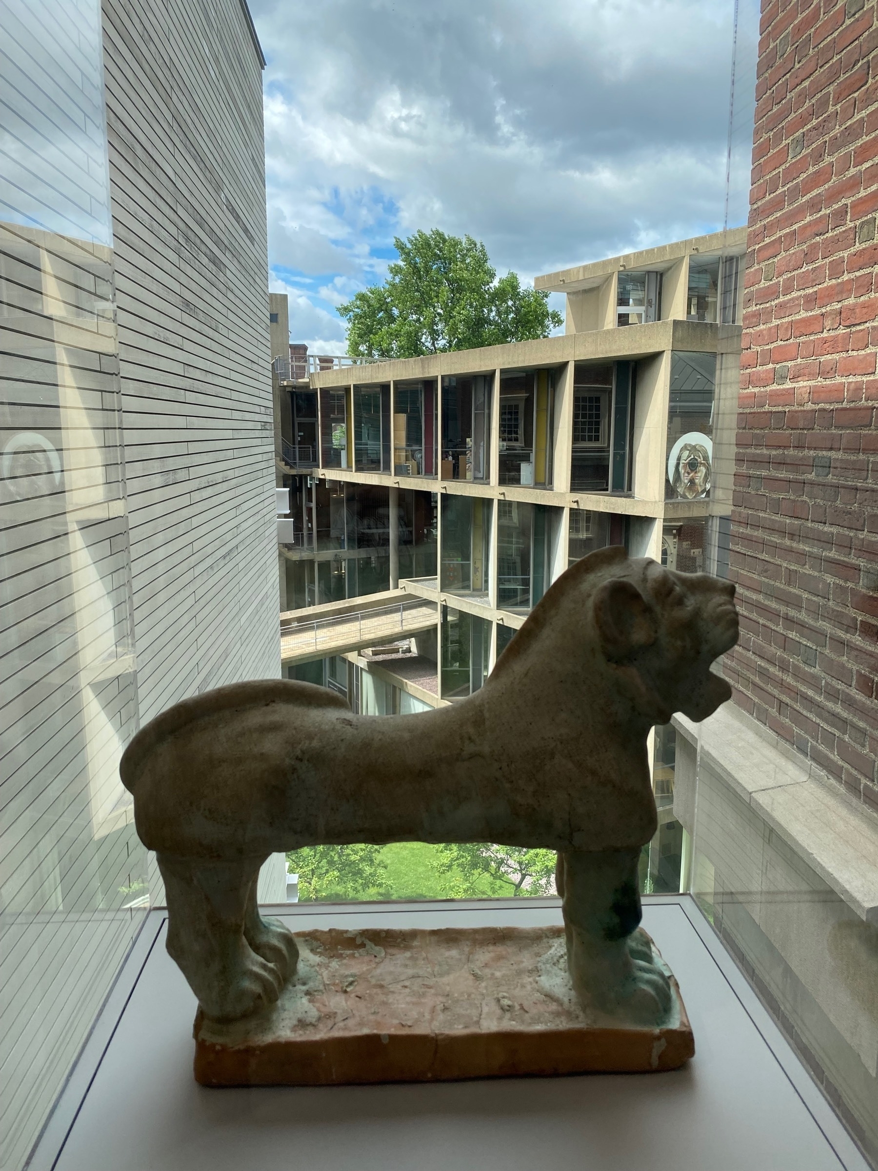 Stone figure of a lion standing in front of a window, with buildings visible in the background.