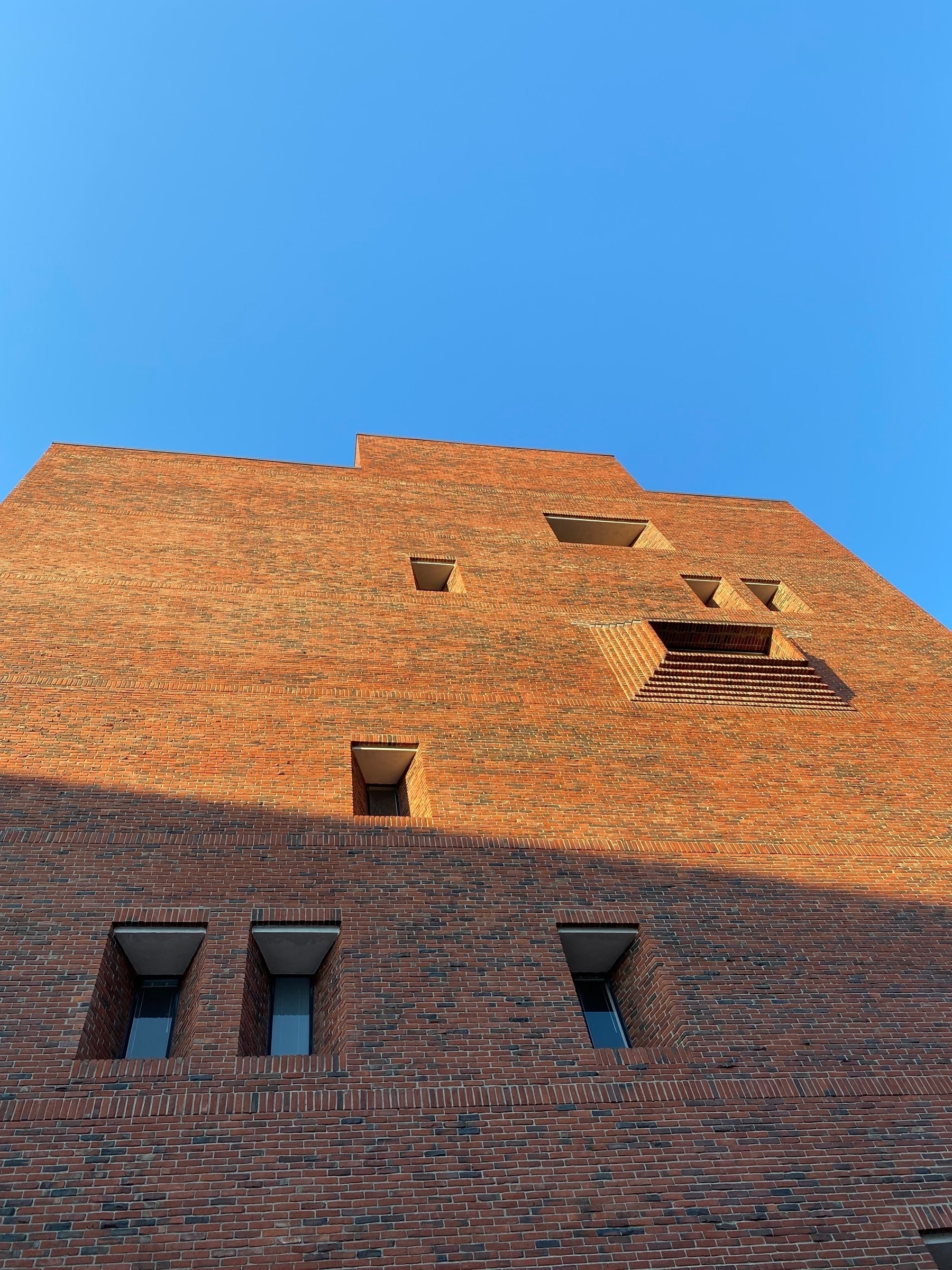 View up the side of a tall brick building, with sunlight on the top and the blue sky behind.