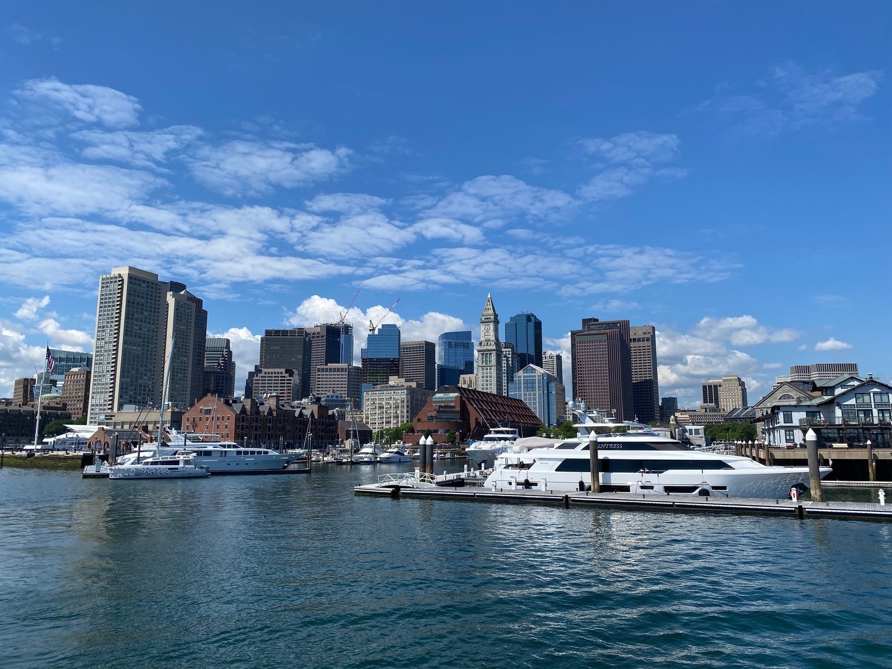 View of Boston's financial district from the harbor, with blue sky above, a white yacht in the foreground, and white clouds behind.