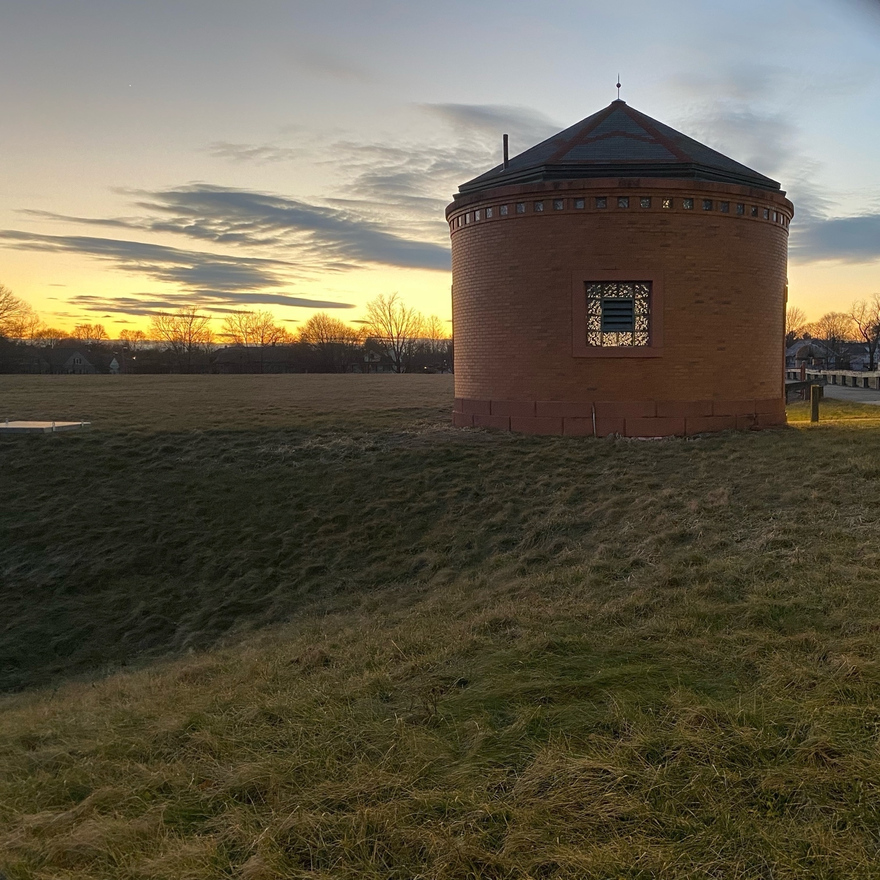 View of a small round brick building with an empty field and sunset behind it.