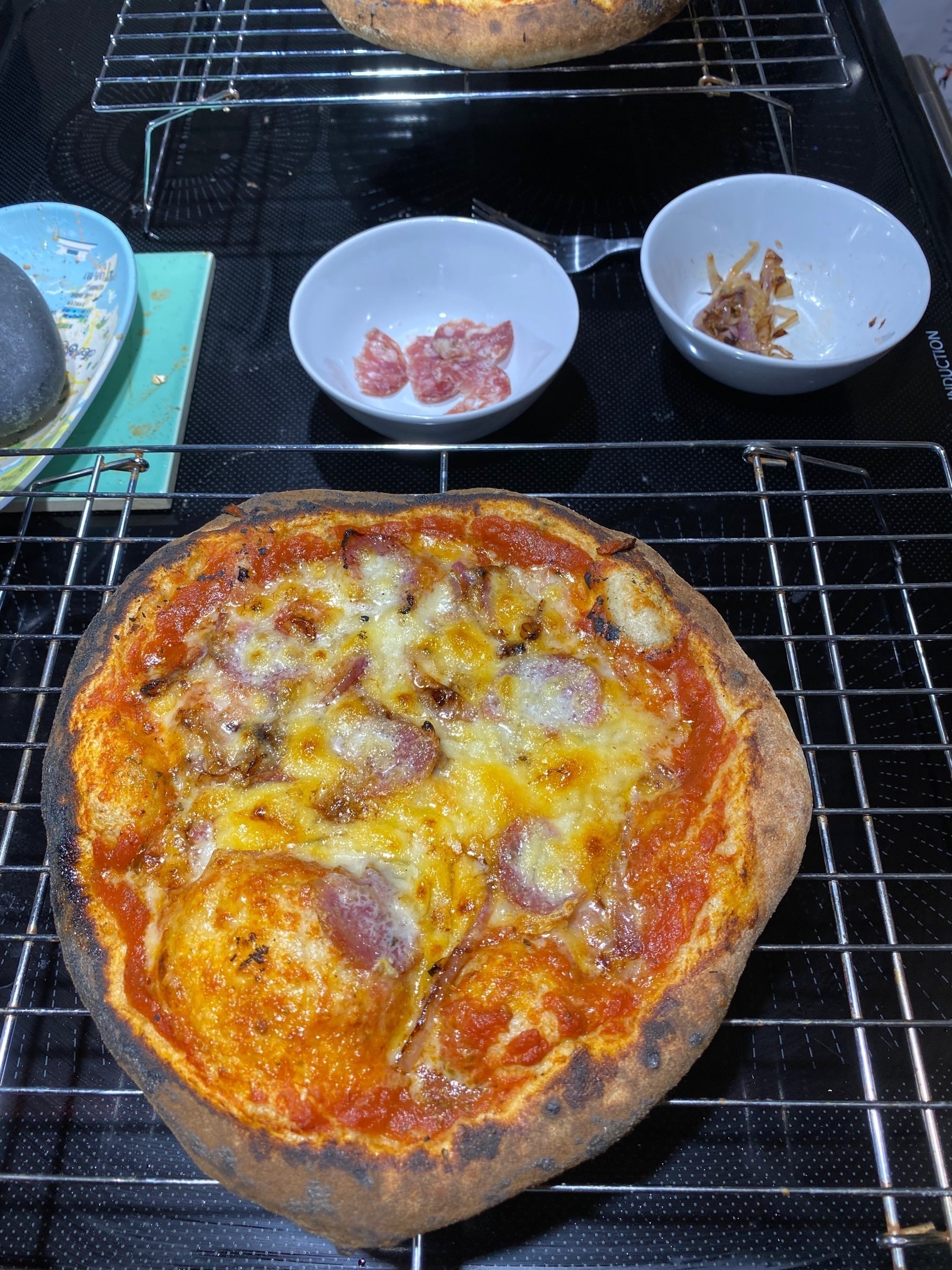 Small pizza on a cooling rack, with small bowls of toppings behind.