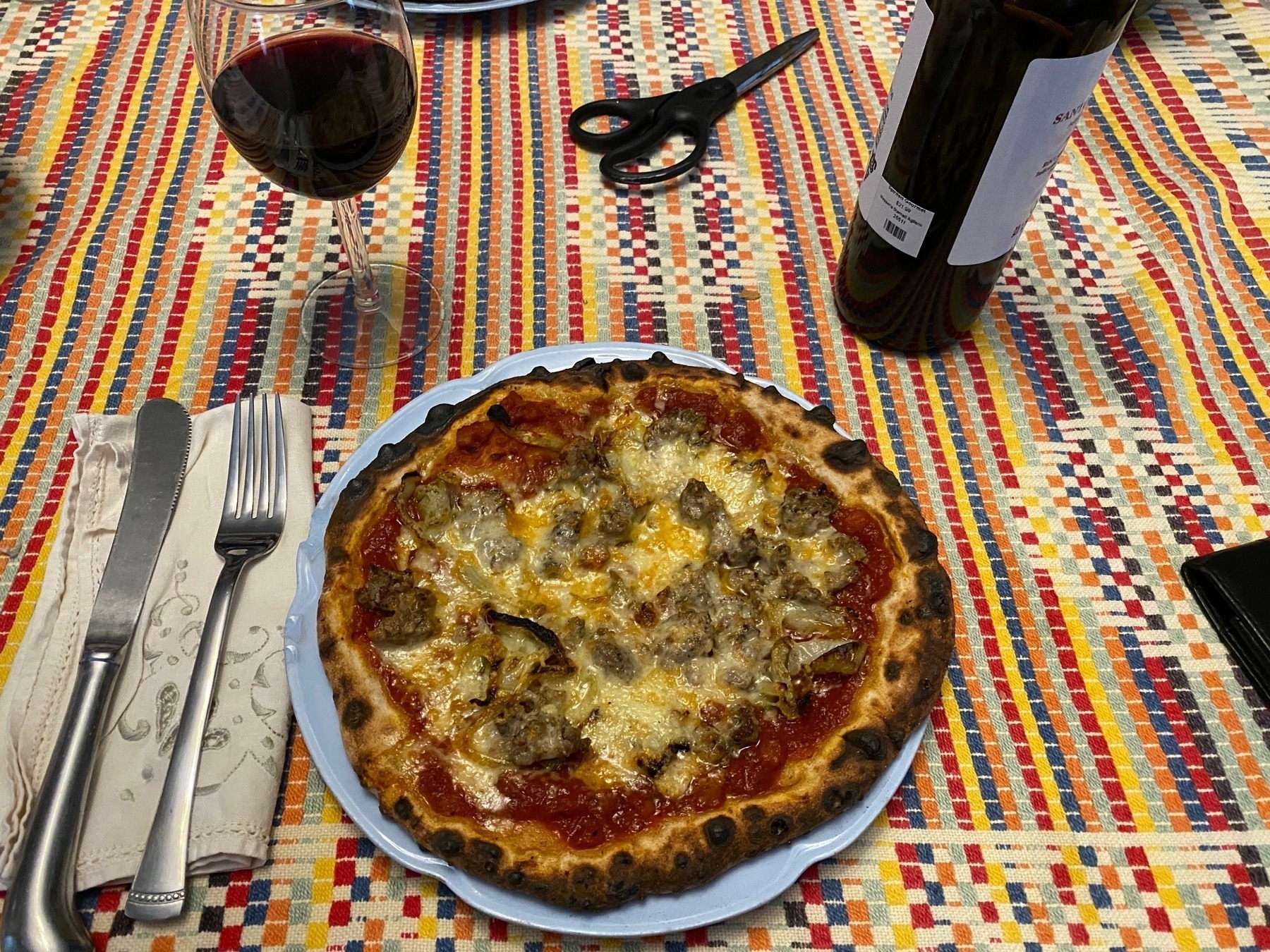 Small pizza on a plate, on a striped tablecloth with utensils, wine glass, and wine bottle nearby.