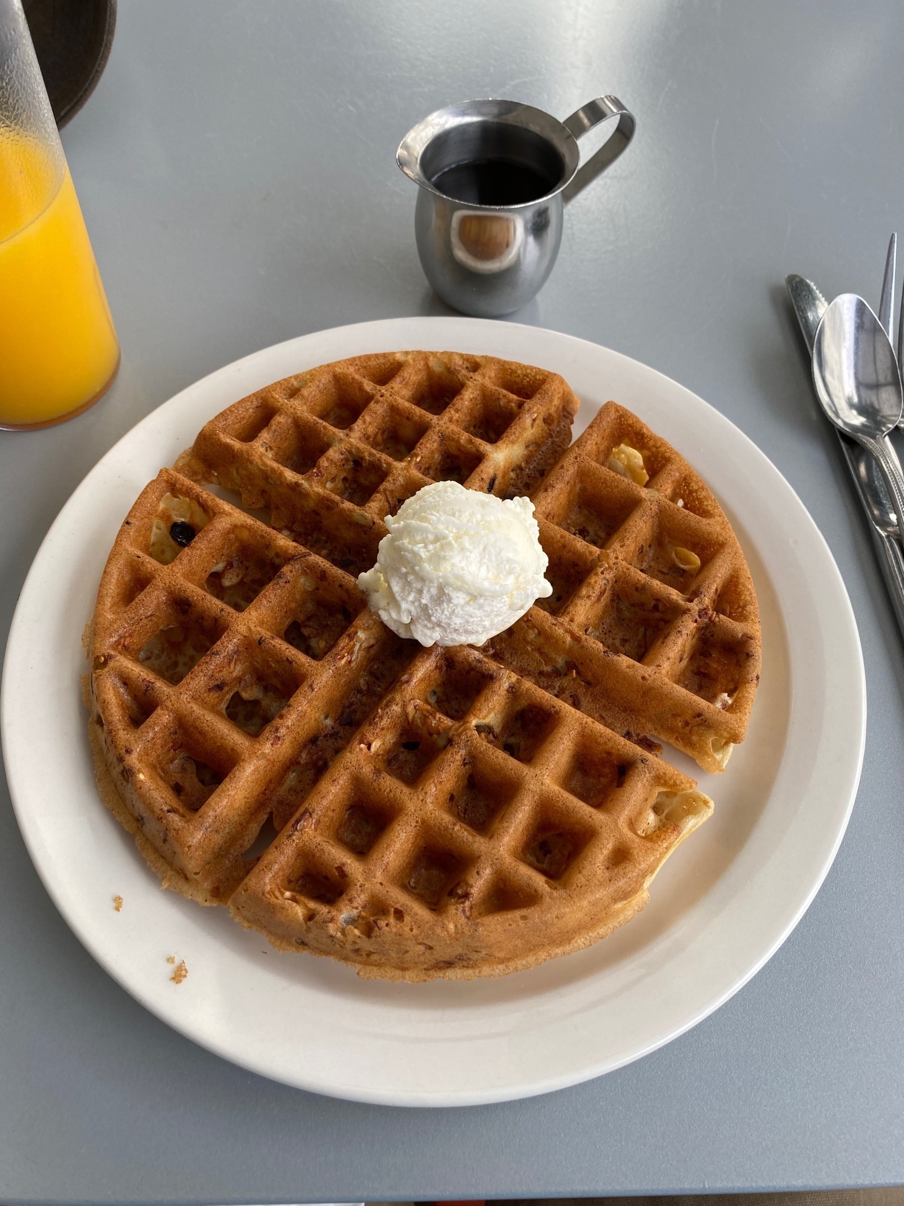 Close up of a round waffle sitting on  a white plate on a gray table, with juice, syrup and silverware nearby.
