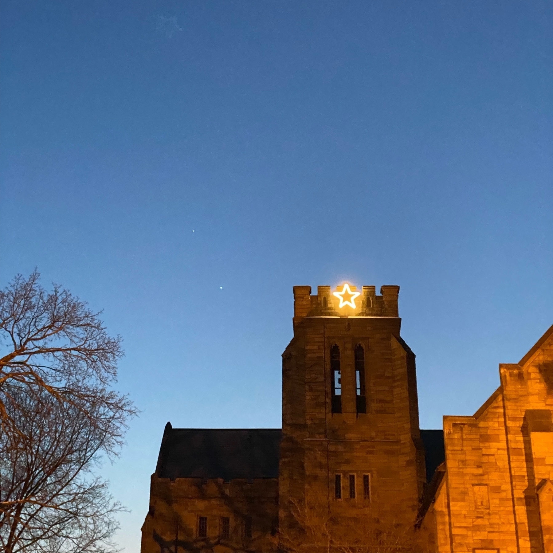 View of a church tower shortly after sunset, with a five pointed illuminated star on the tower and Venus and Jupiter just to the left in the dark blue sky.