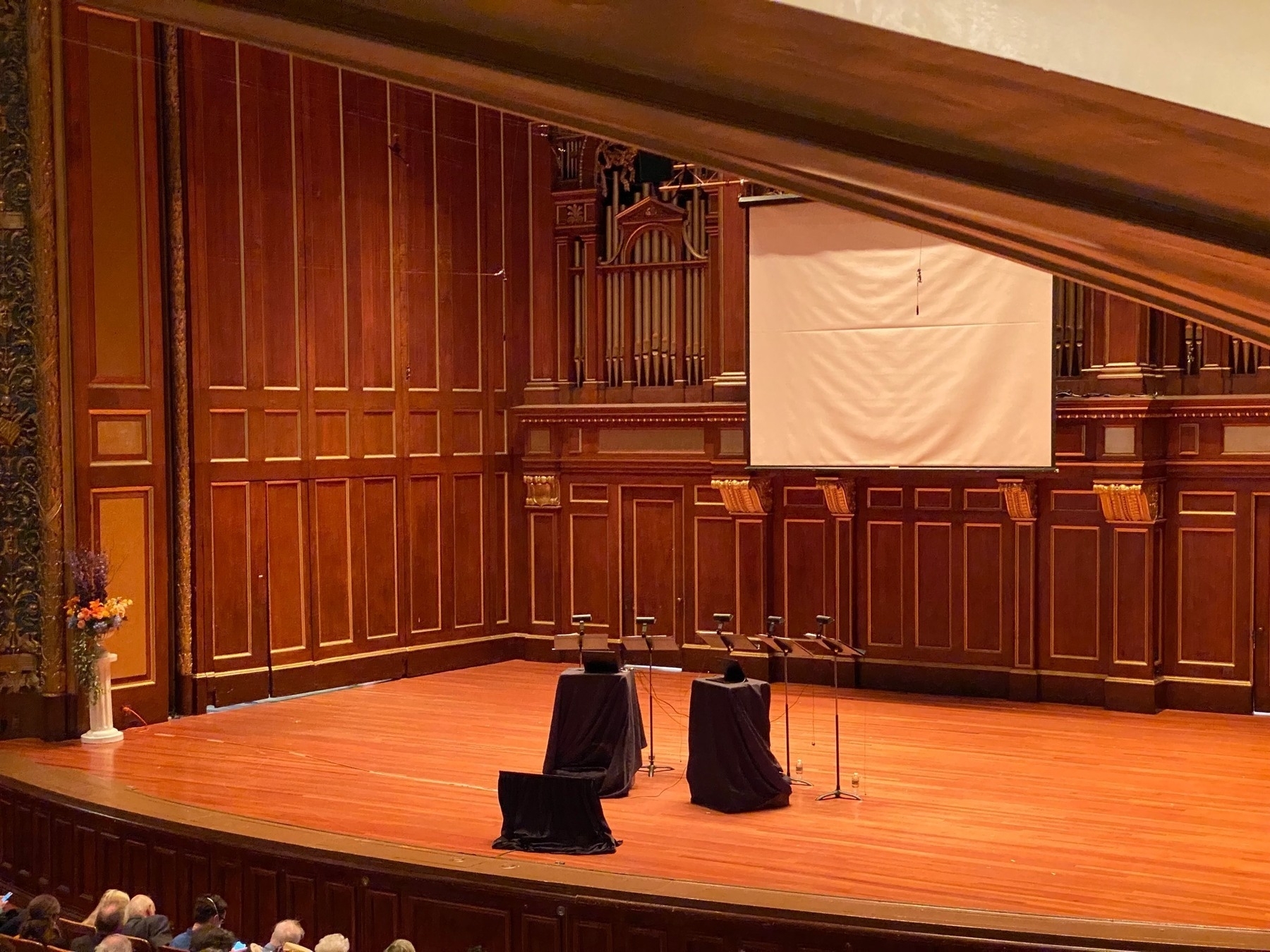 Bare stage with music stands and black boxes center stage, elaborate paneled walls behind, and a dumpy looking pull down screen on the wall.
