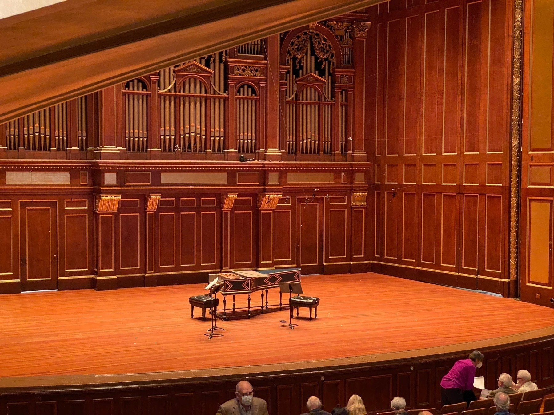 Bare wooden stage with a harpsichord and music stands in the center, with wood panels and a large organ built into the back wall.