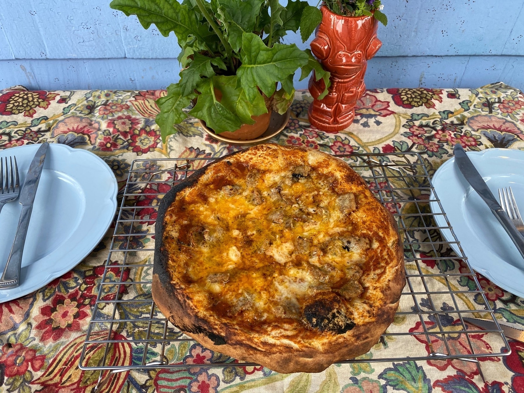 Small pizza on a cooling rack, sitting on a table with a floral cloth, blue plates, and flower vases.