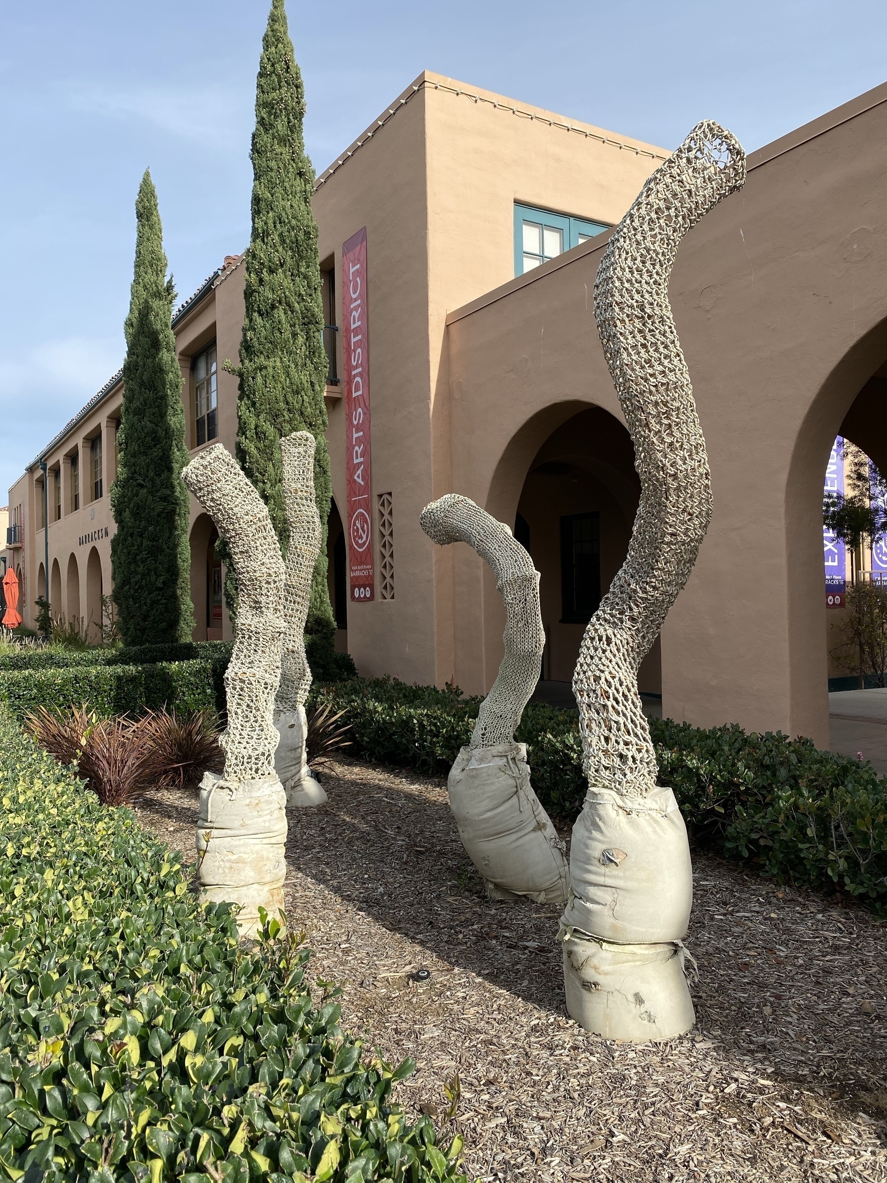 White woven tube sculptures in a hedge garden in front of a light brown building.