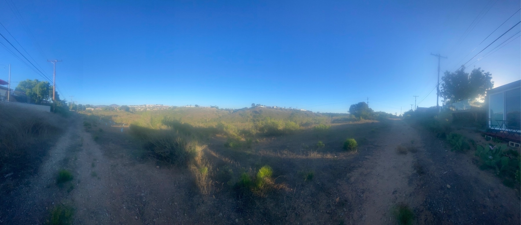 Panoramic view from the rim of a scrub canyon with houses lining the far side and a blue sky above.