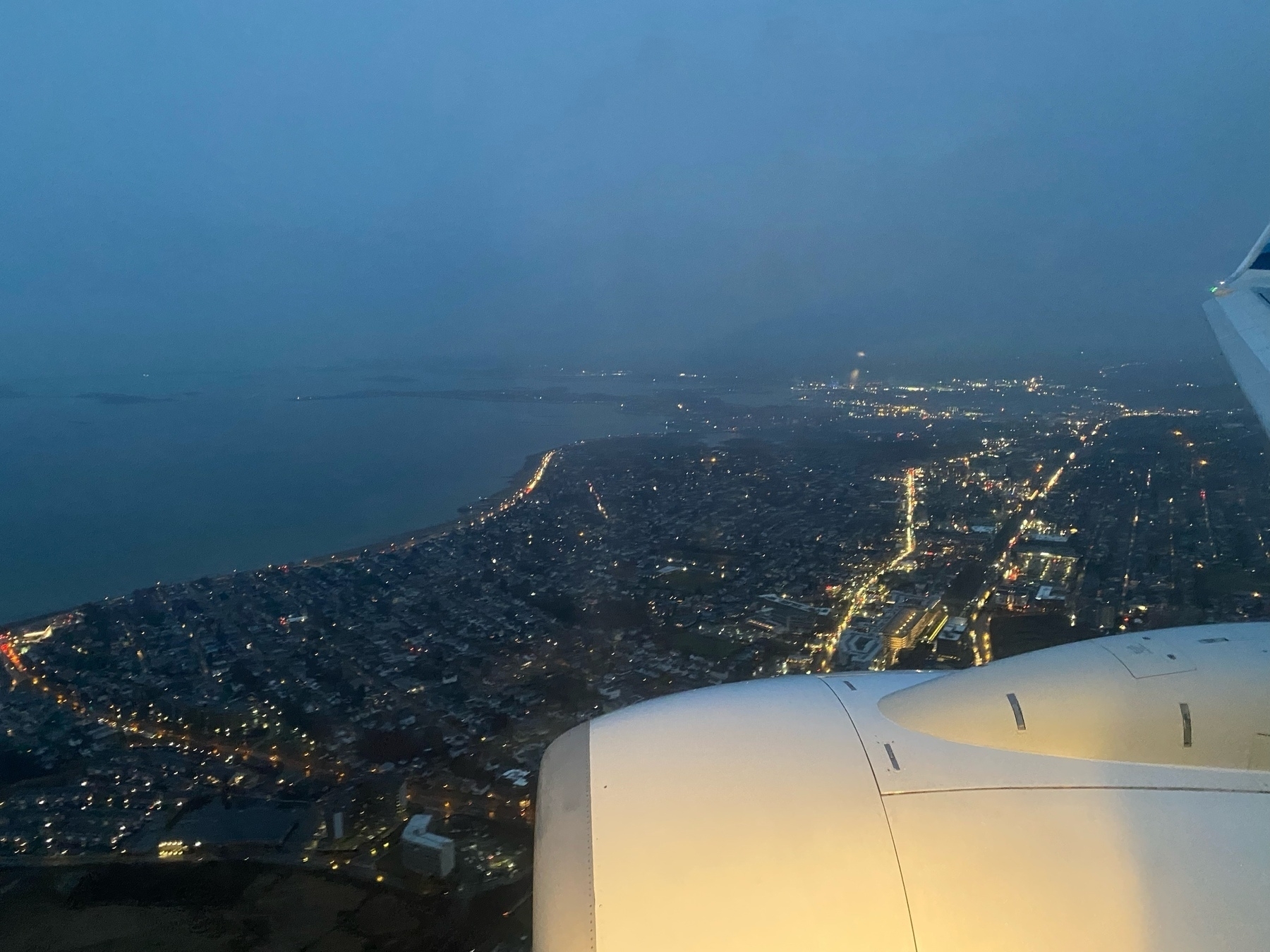 View out a plane window of the shoreline in the evening with the lights of streets and houses below.