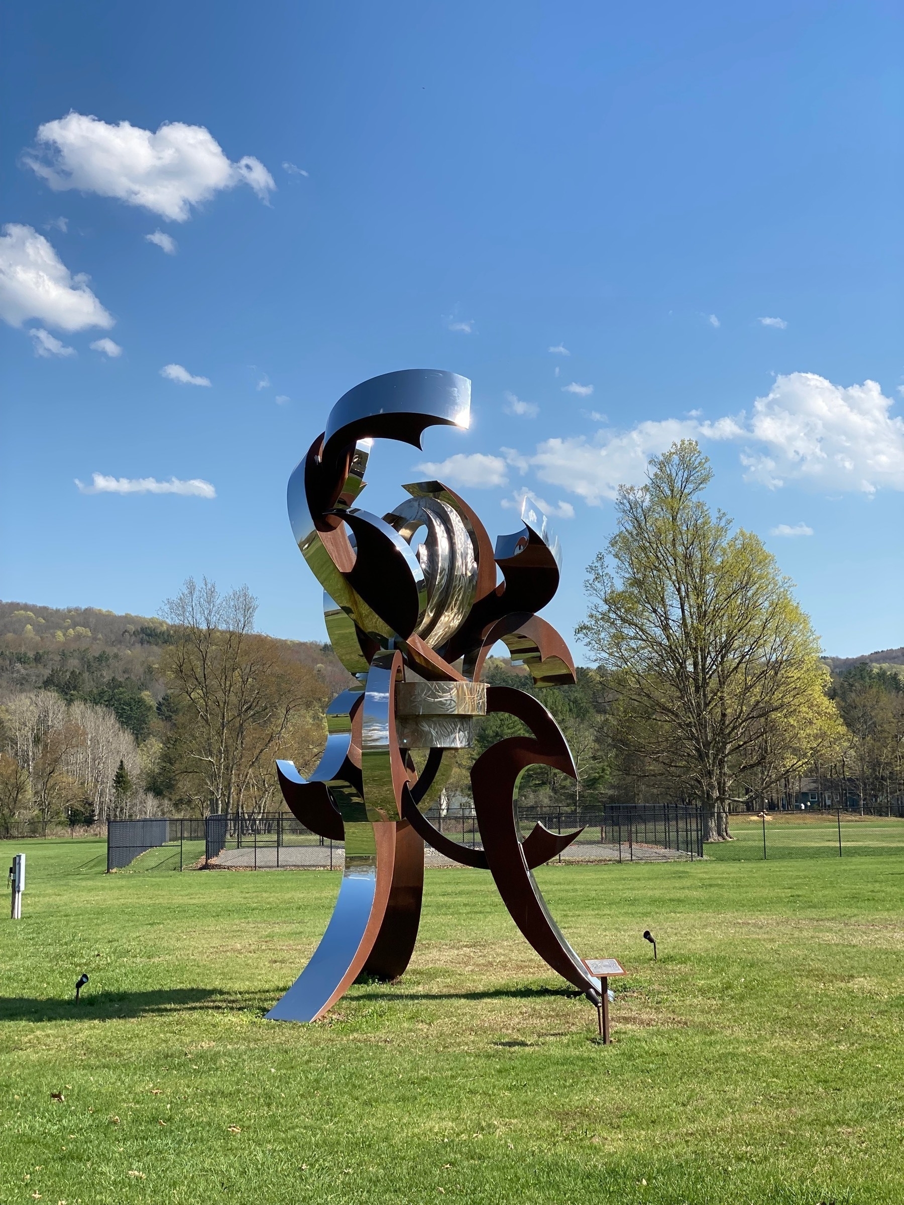 Metal sculpture in a grassy field with trees and hills in the background, a blue sky above.
