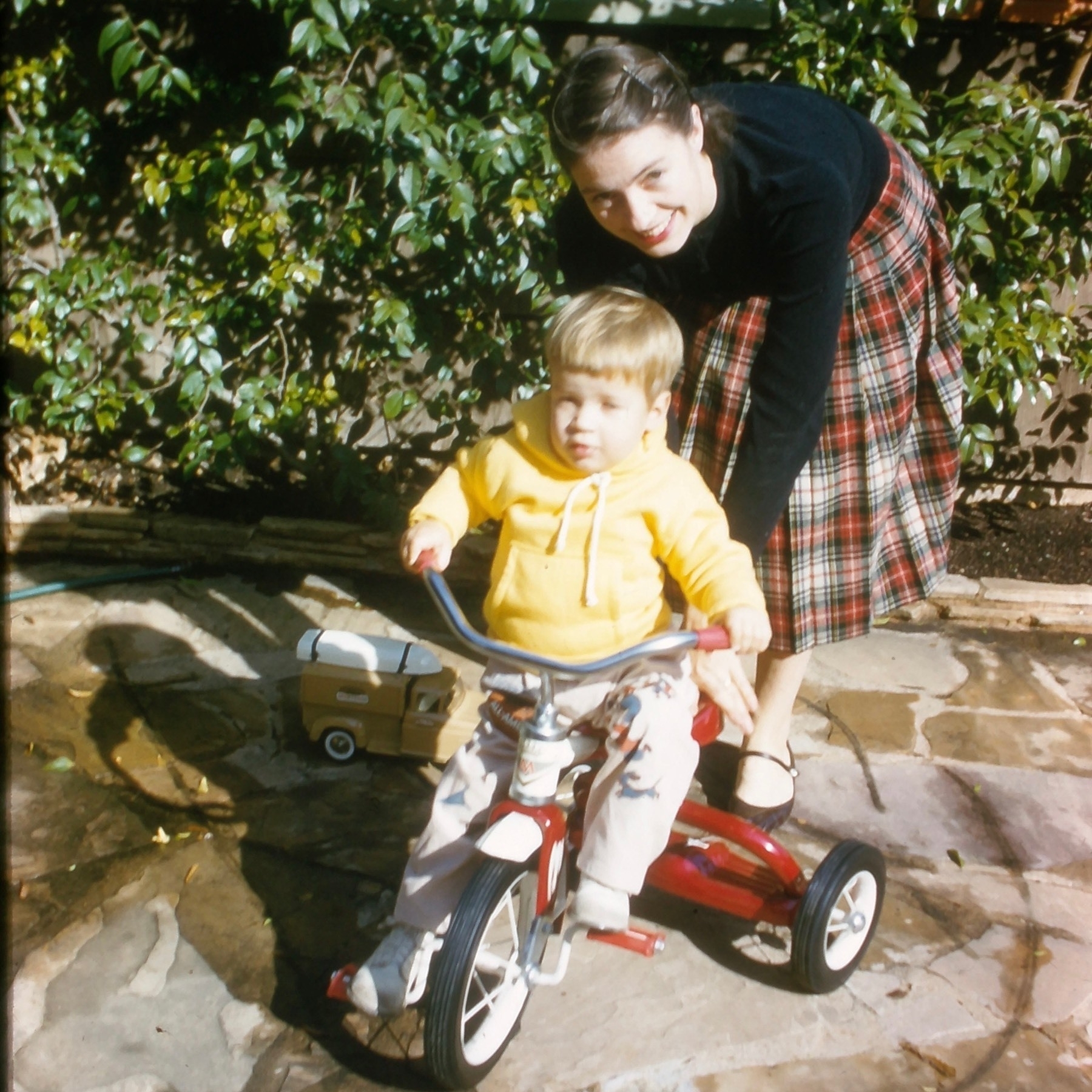 Toddler on a tricycle with his mother leaning behind.