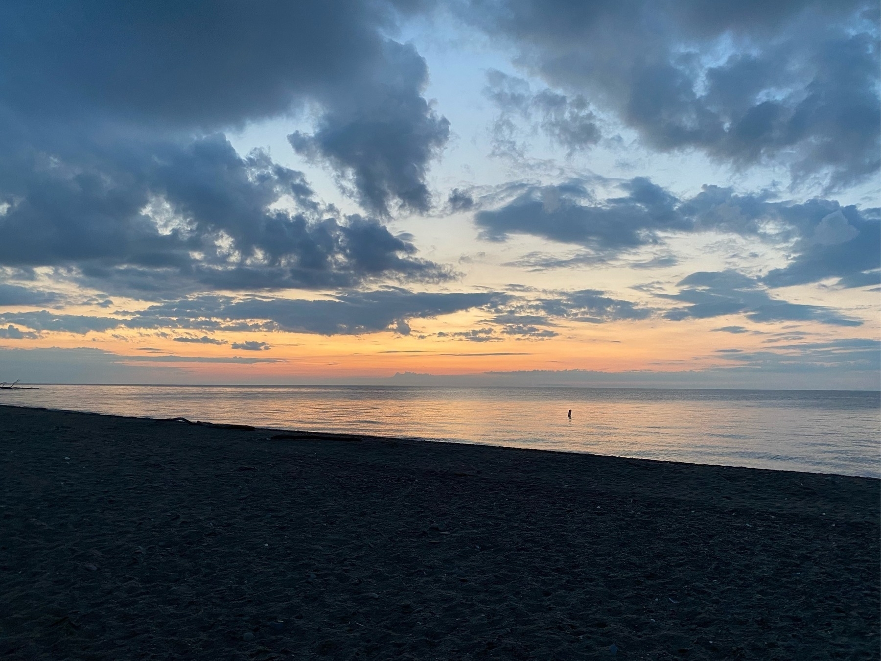 View of sunset on a large lake, with many dark clouds above, a line of orange at the horizon where the sun has just set, and a wedge of black beach in the foreground.