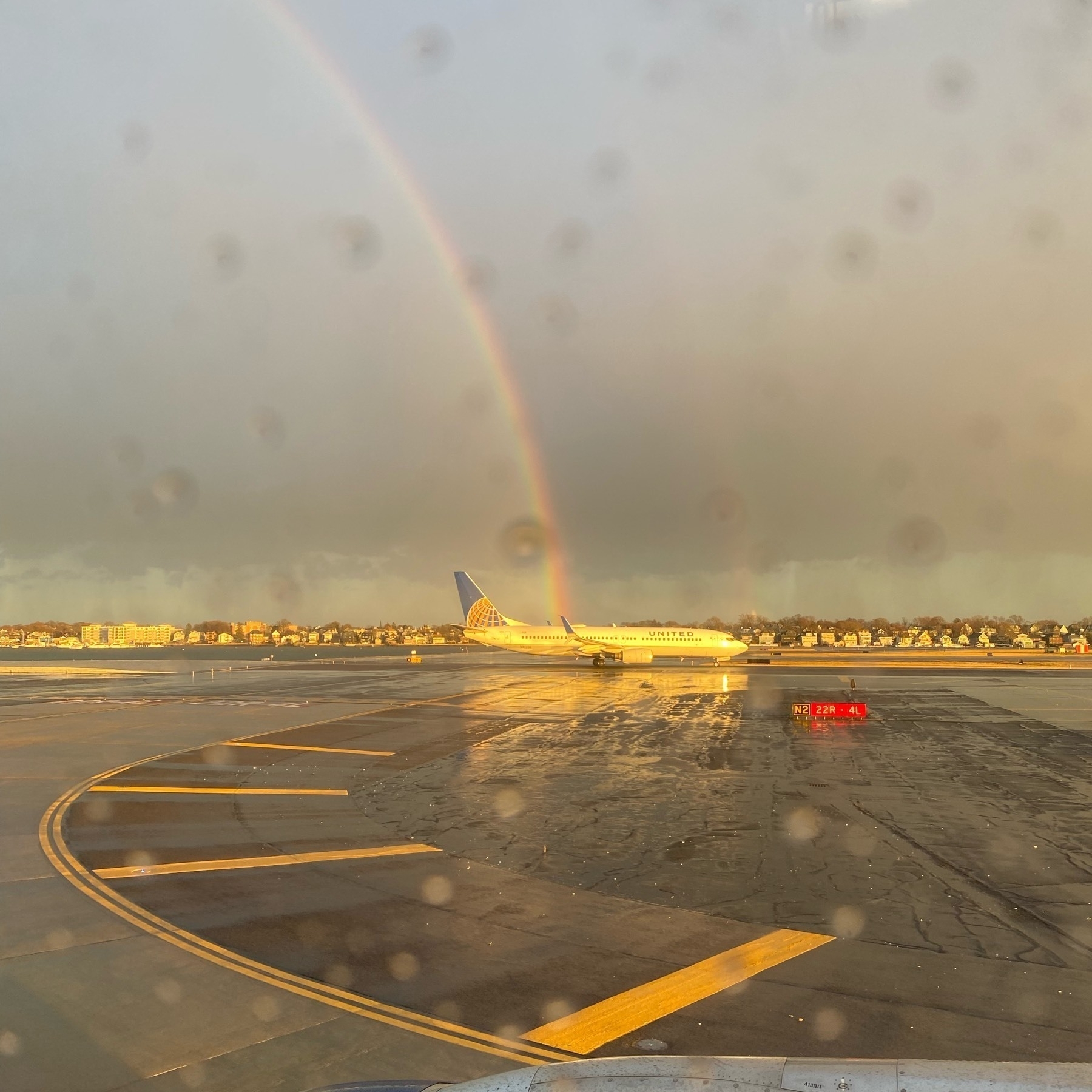 View out an airplane window of another plane on a runway lit by the setting sun with half a rainbow in the sky reaching the ground just behind the other plane.