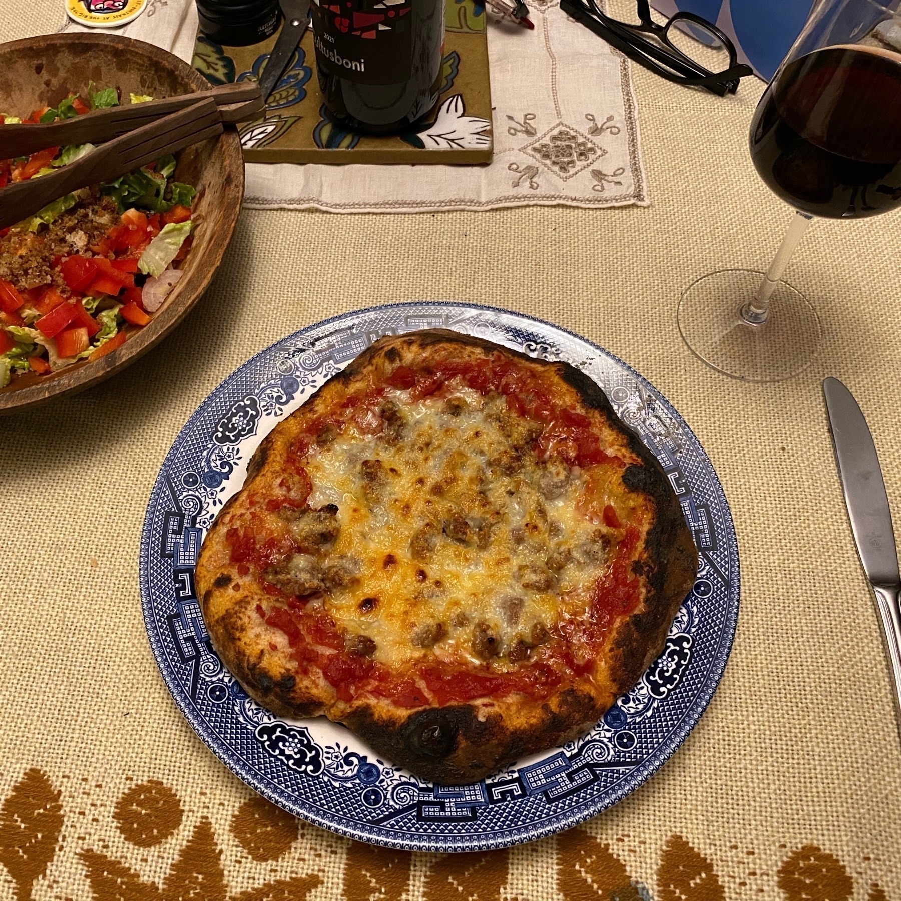 Small pizza on a blue plate, sitting on a table with a light brown tablecloth, salad, wine and utensils in the background.