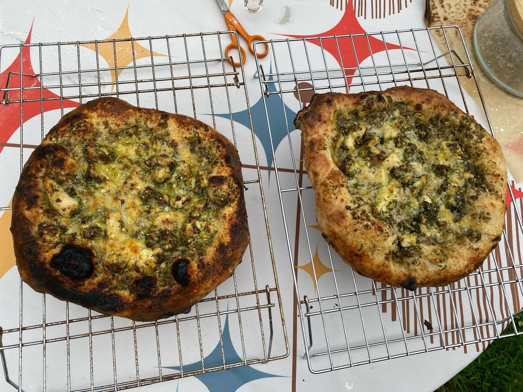 Two small chicken pesto pizzas on cooling racks, on a table with a white cover with various color starbursts.