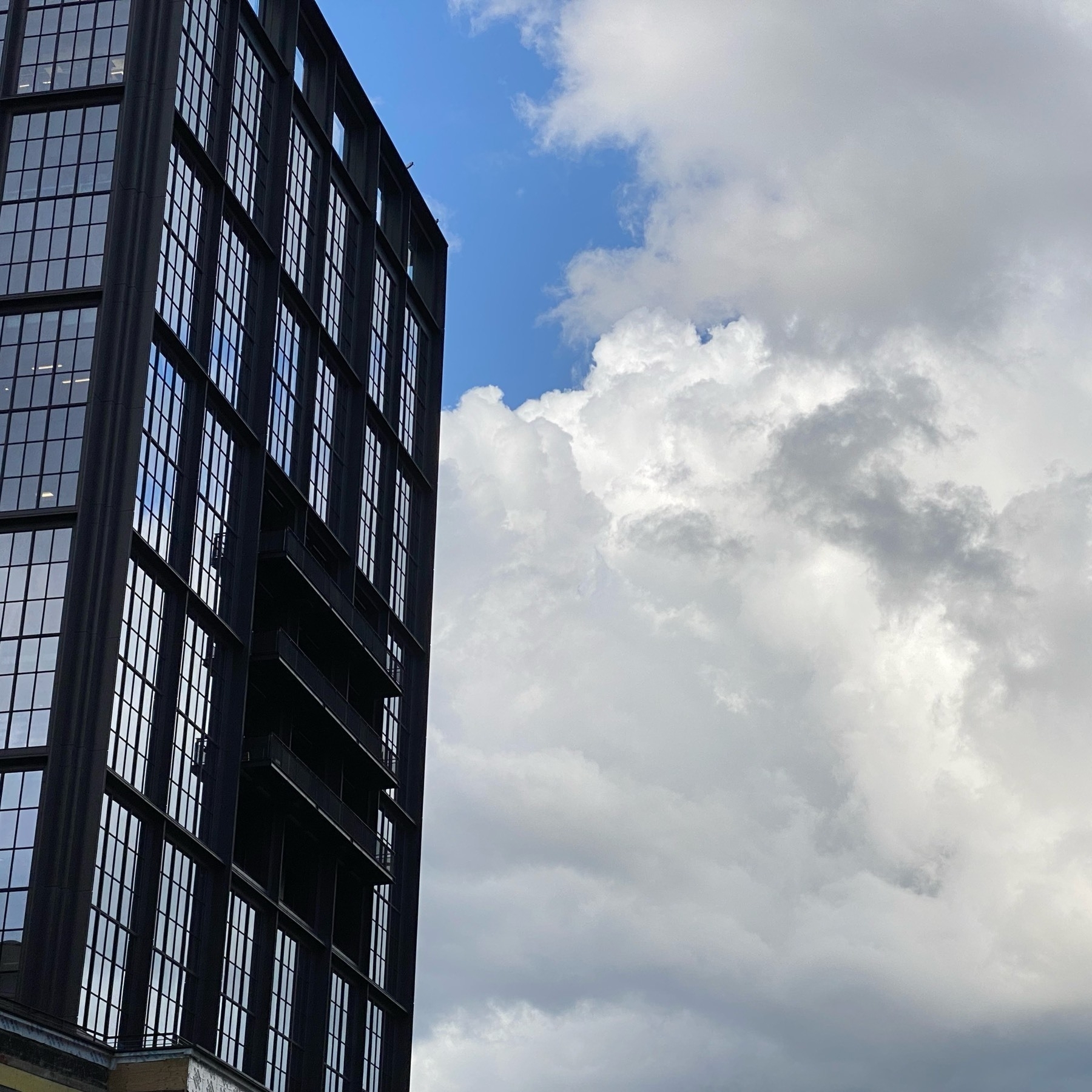 View of puffy white clouds against a blue sky, reflected in the windows of a building on the left with dark black frames and structural elements. 