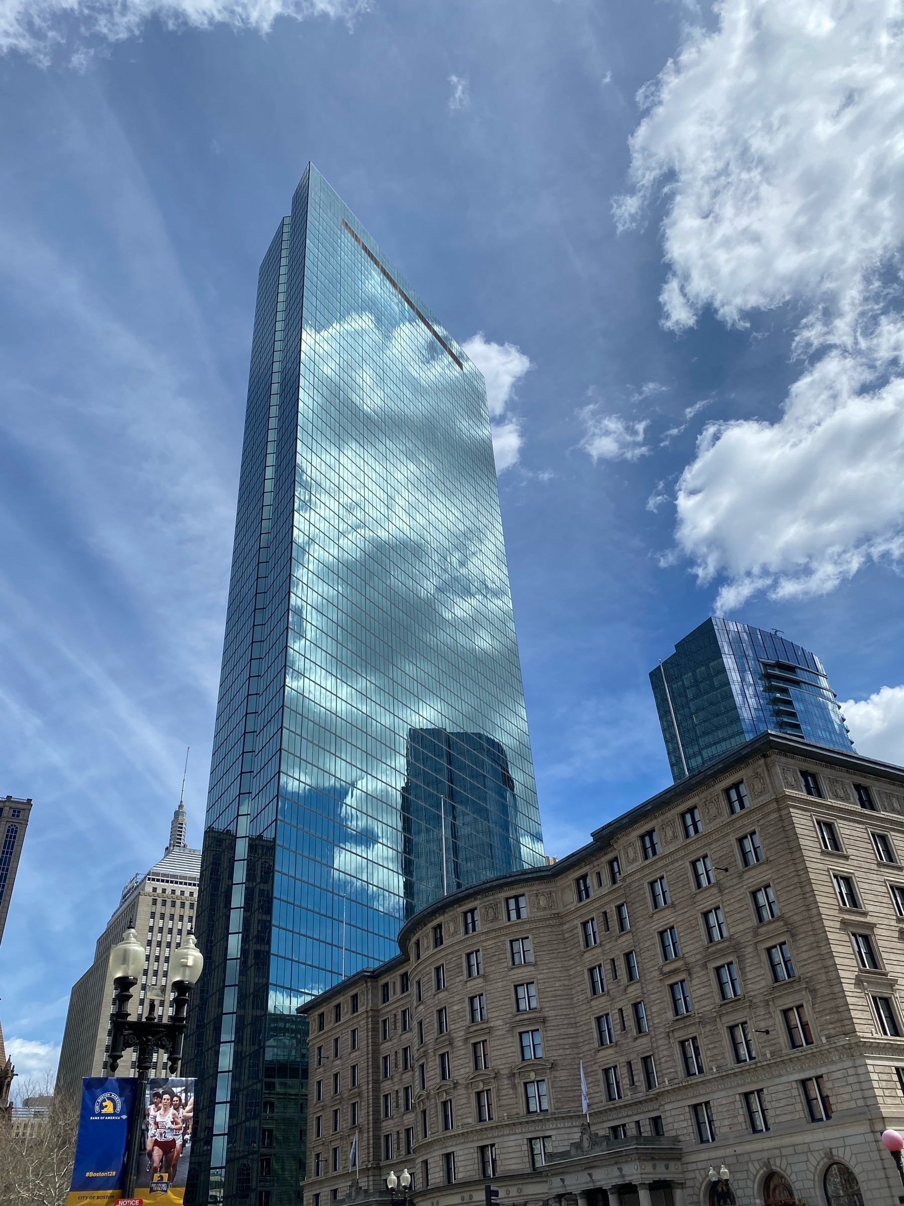 View of the Hancock skyscraper in Copley Square, Boston, with backlit clouds reflected from the windows and the blue sky behind.