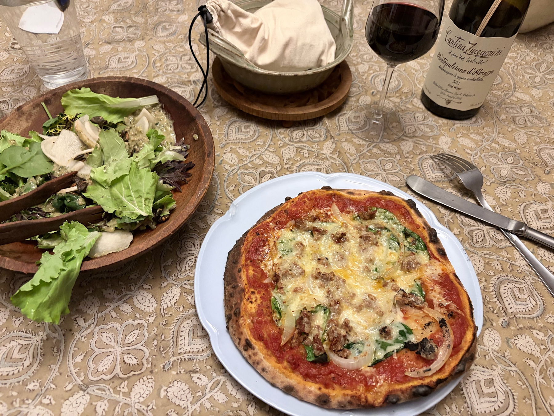 A small pizza on a light blue plate on a brown paisley tablecloth, next to a wooden salad bowl, a bottle of wine, and knife and fork. 