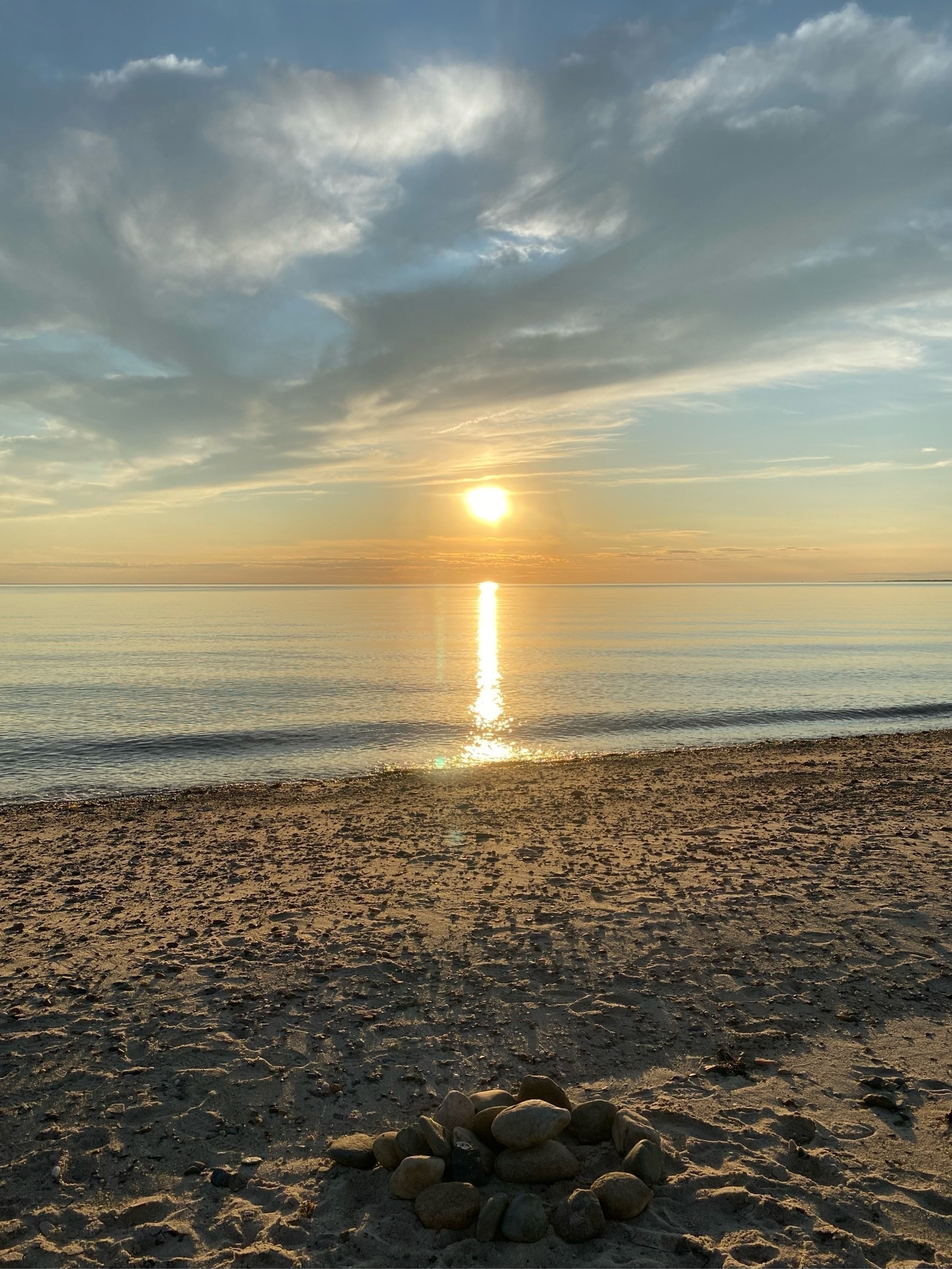 View of an orange sunset over calm water, the sun just above the horizon, a pale blue sky with wispy white clouds above, a dark stony beach below.