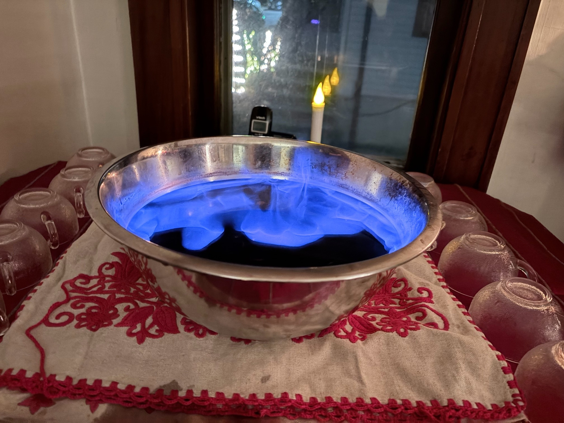 Blue flames on the surface of a metal bowl of punch, lines of punch glasses on either side, a dark window in the background reflecting a small electric candle.