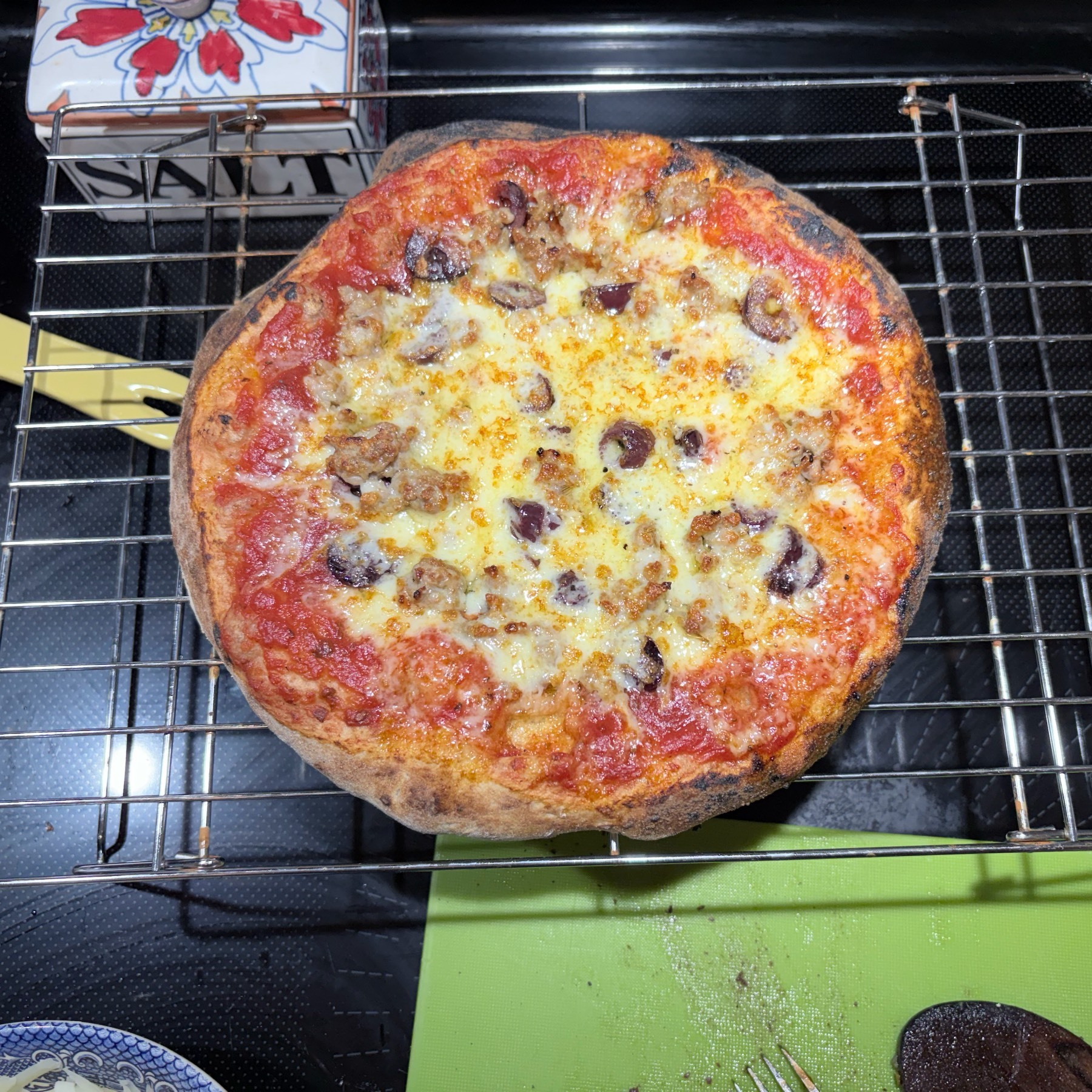 Small pizza on a metal wire cooling rack, sitting on a black glass stove top, with part of a green cutting board and a white salt box nearby. 