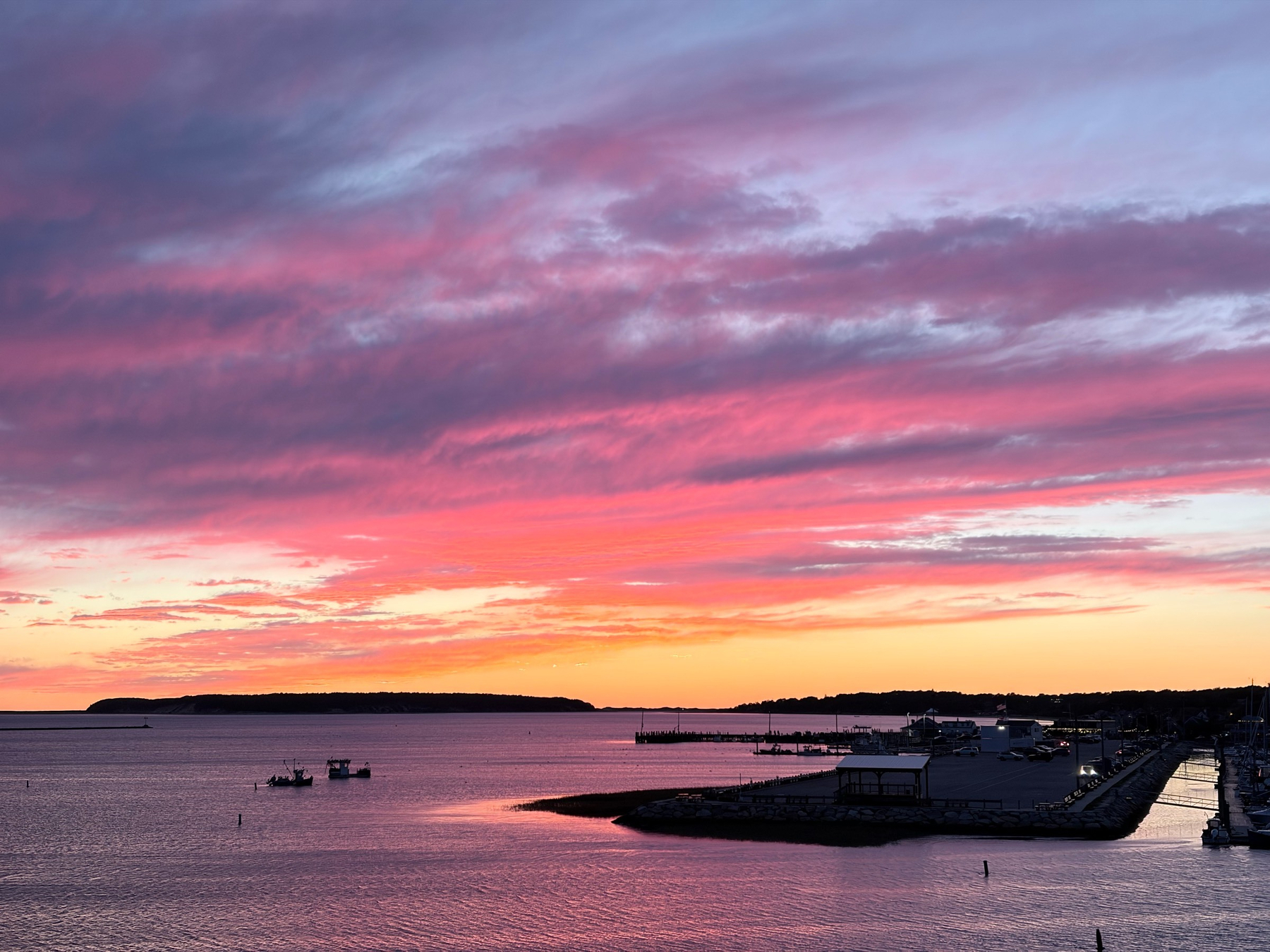 View of sunset over a harbor, with a dark pier below and several small boats adjacent.