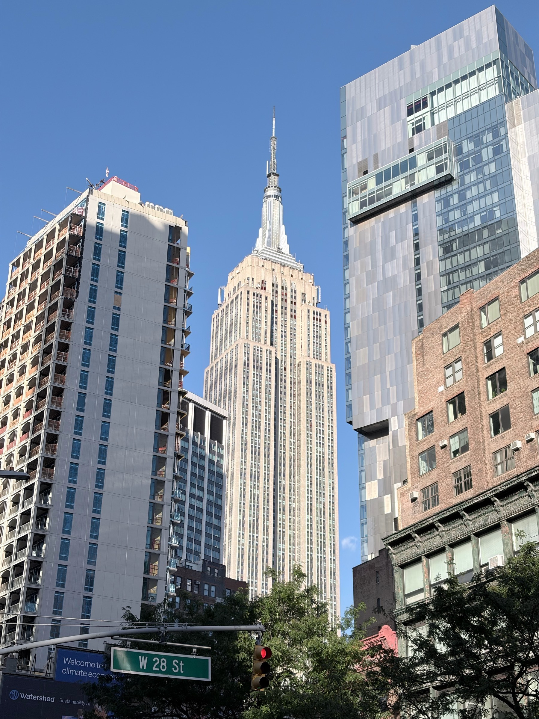 View of the Empire State Building from 28th Street, the building framed on either side by high rise apartment towers, blue sky beyond. 