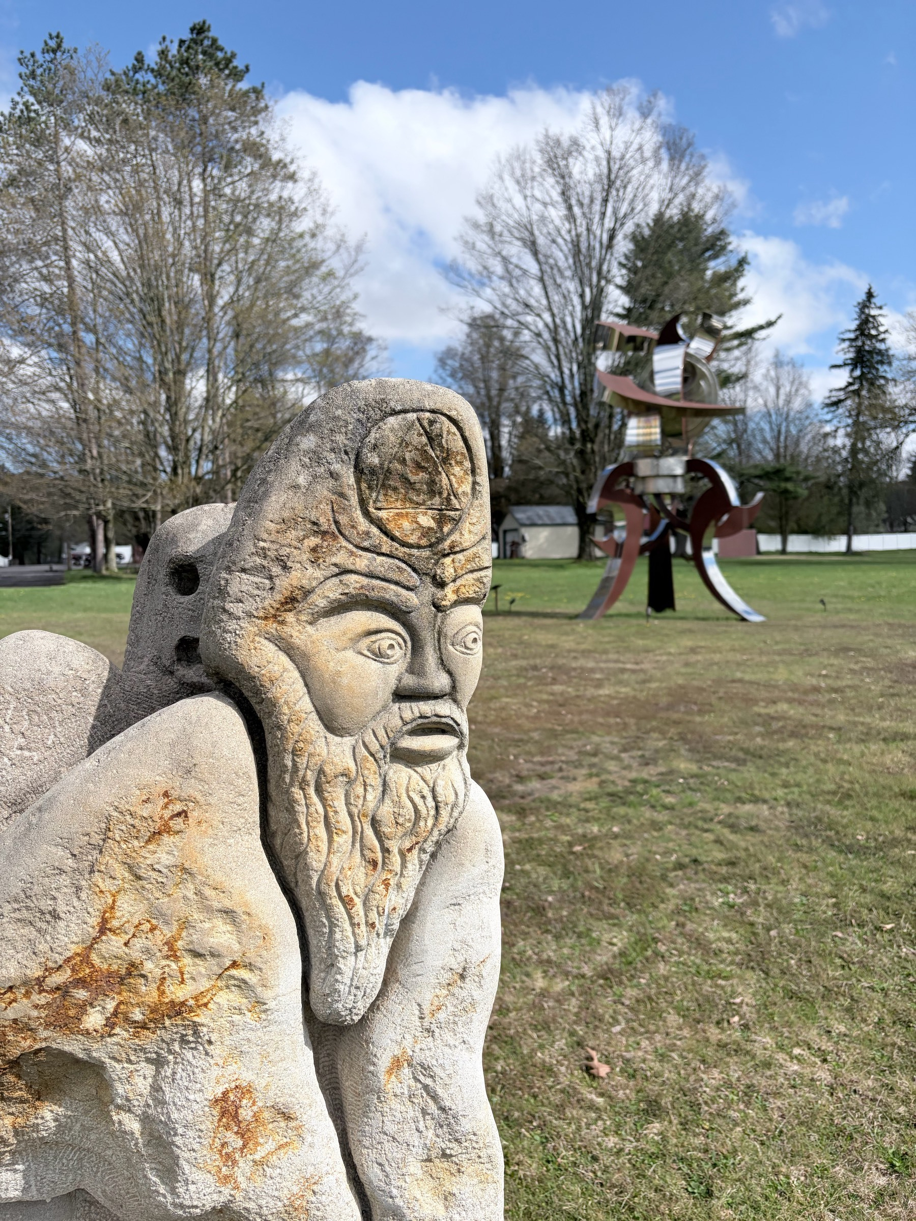 A stone sculpture in the left foreground of a beaded man, with a large metal sculpture in the right background sitting on a lawn, with trees and white clouds against a blue sky behind.