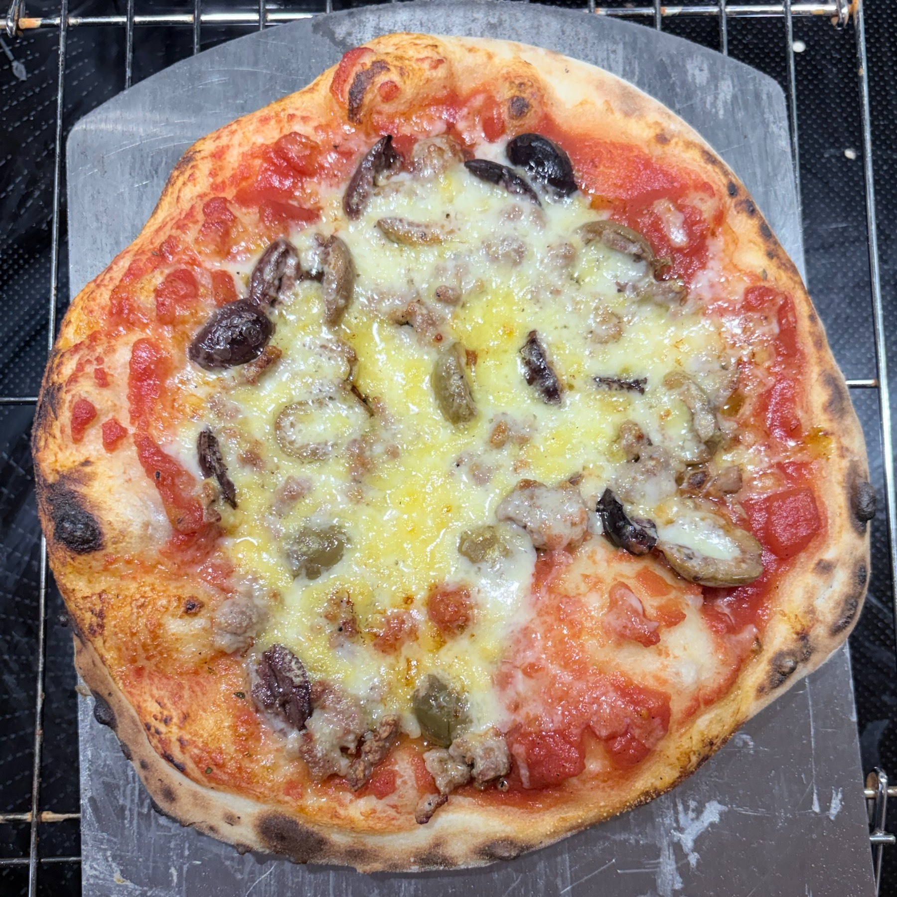 Small pizza sitting on a silver peel, which is sitting on a wire cooling rack, which is sitting on a black glass stove top.