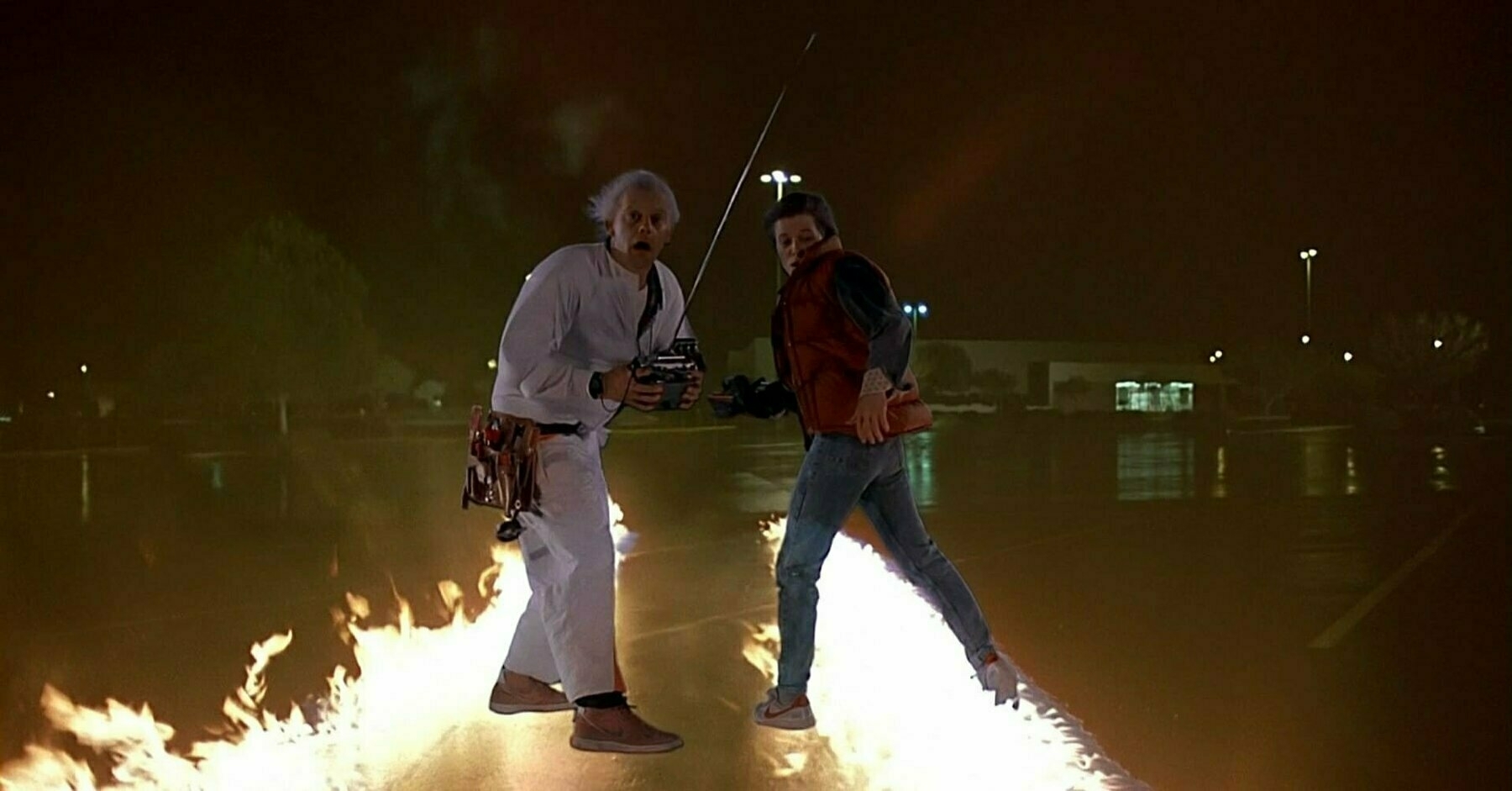 A white-haired scientist and his young protégé stand atop a pair of flaming tire tracks in the middle of a dark parking lot.