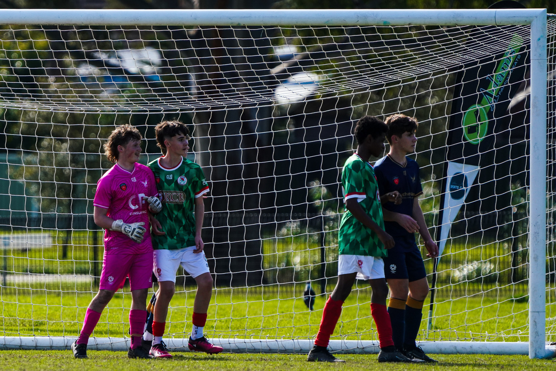 Four soccer players, including a goalie in pink, stand in front of a goalpost during a game.