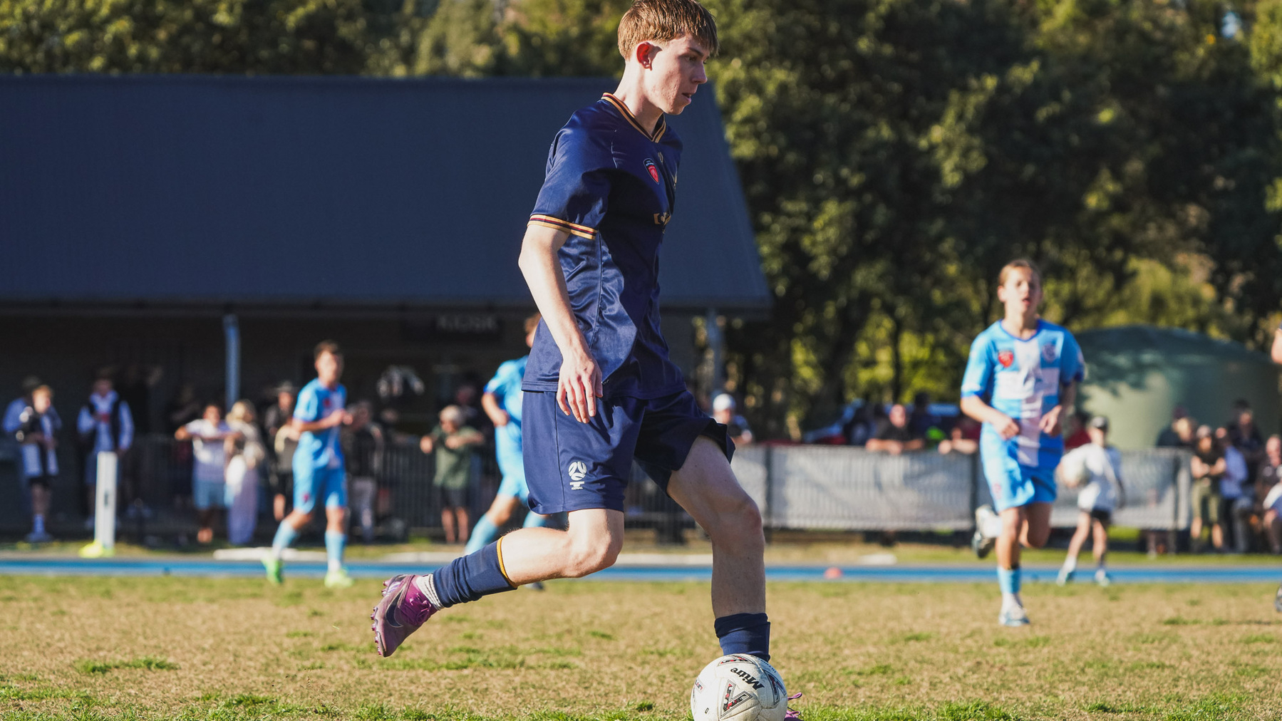 A soccer player in a dark blue uniform is dribbling the ball on a field while other players and spectators are visible in the background.