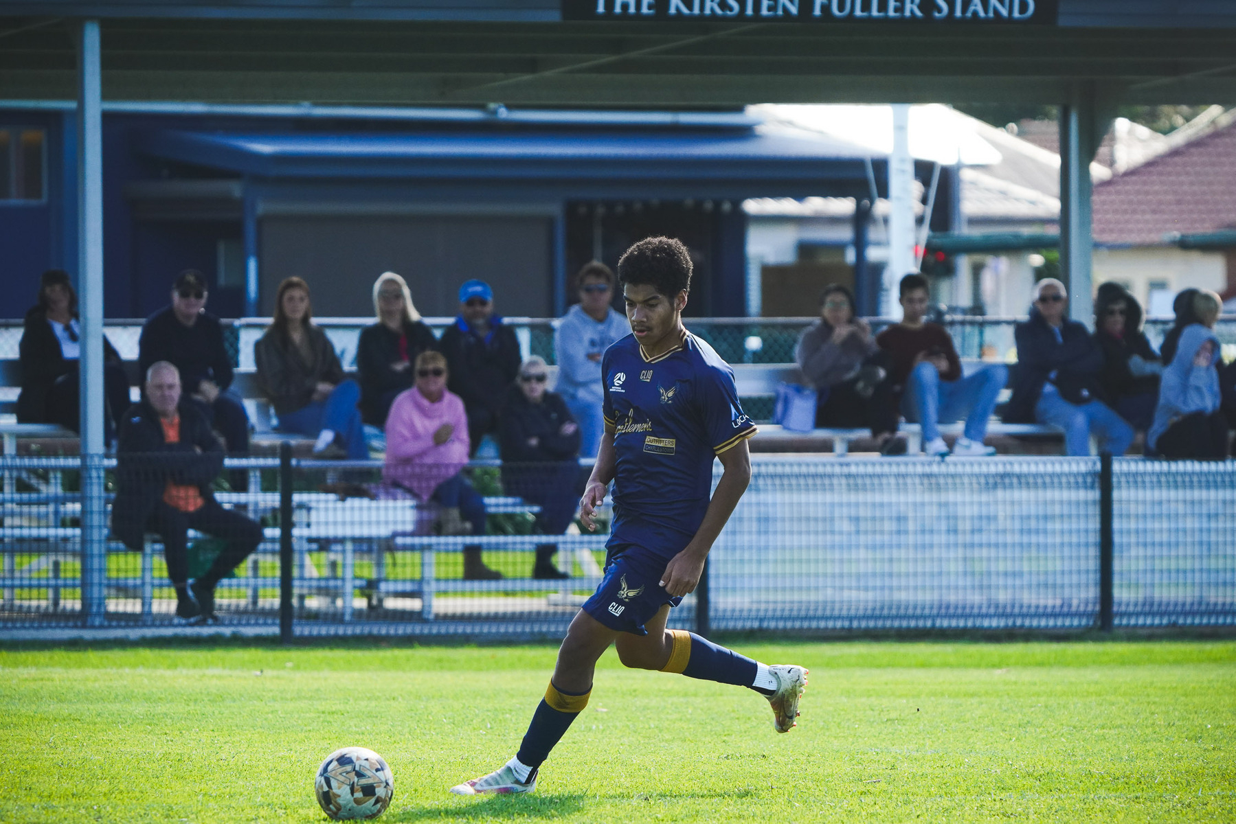 A soccer player in a dark blue jersey is dribbling the ball on a grassy field, with spectators seated in the stands behind him.