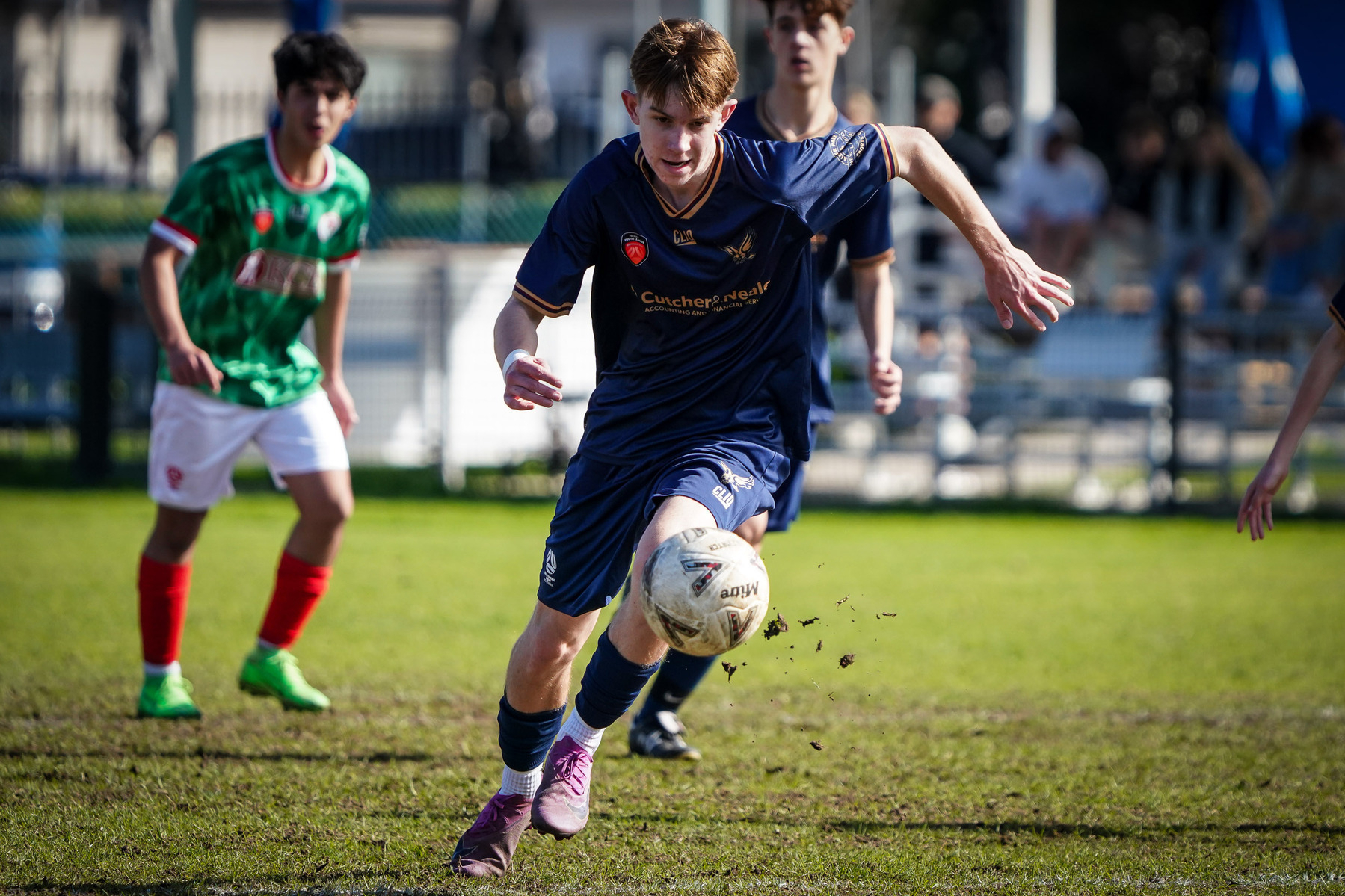 A soccer player in blue appears focused as he runs towards the moving ball during a match, with other players and spectators in the background.