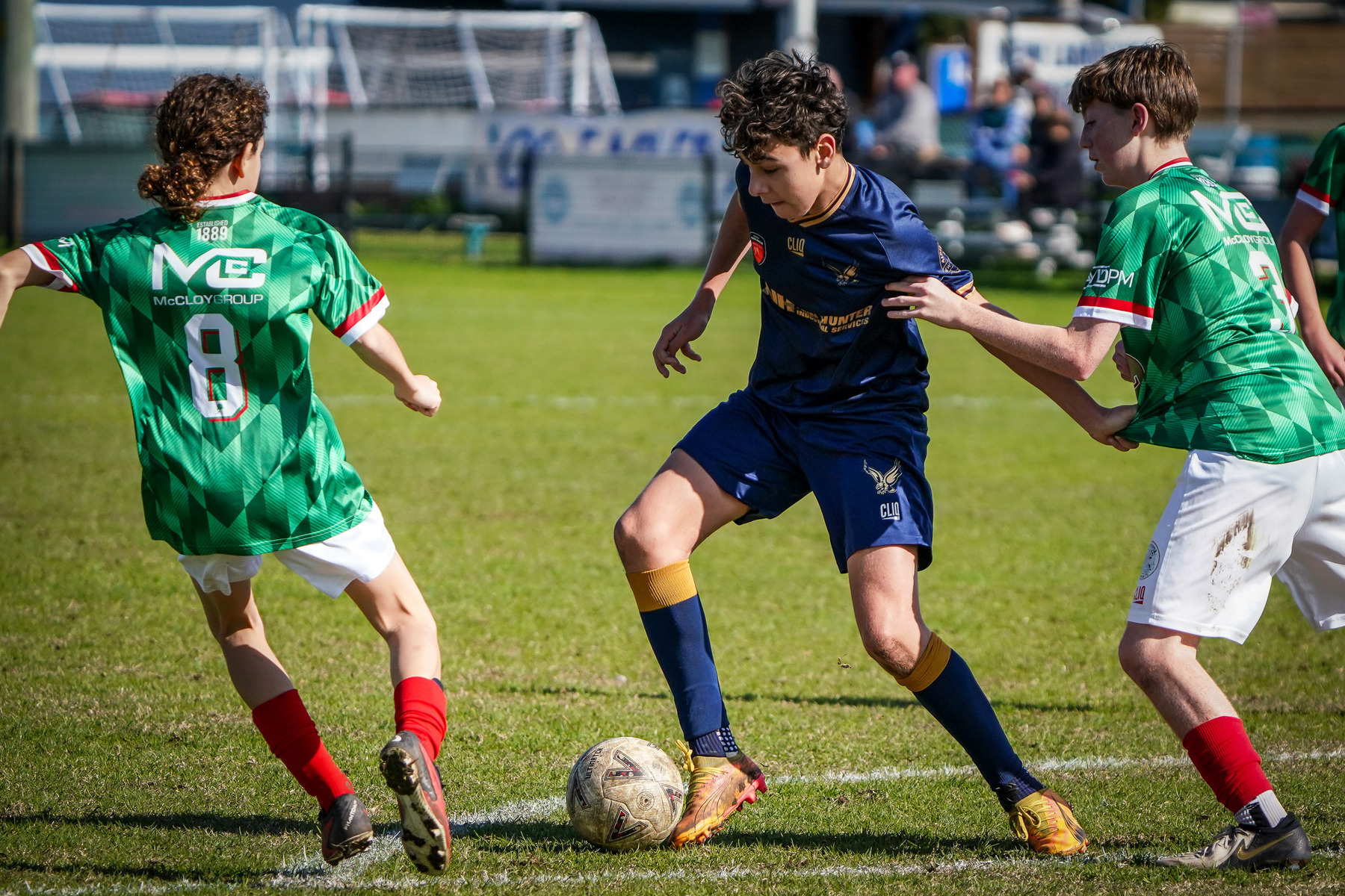 Three young soccer players, two in green jerseys and one in blue, are vying for control of the ball on a grassy field.